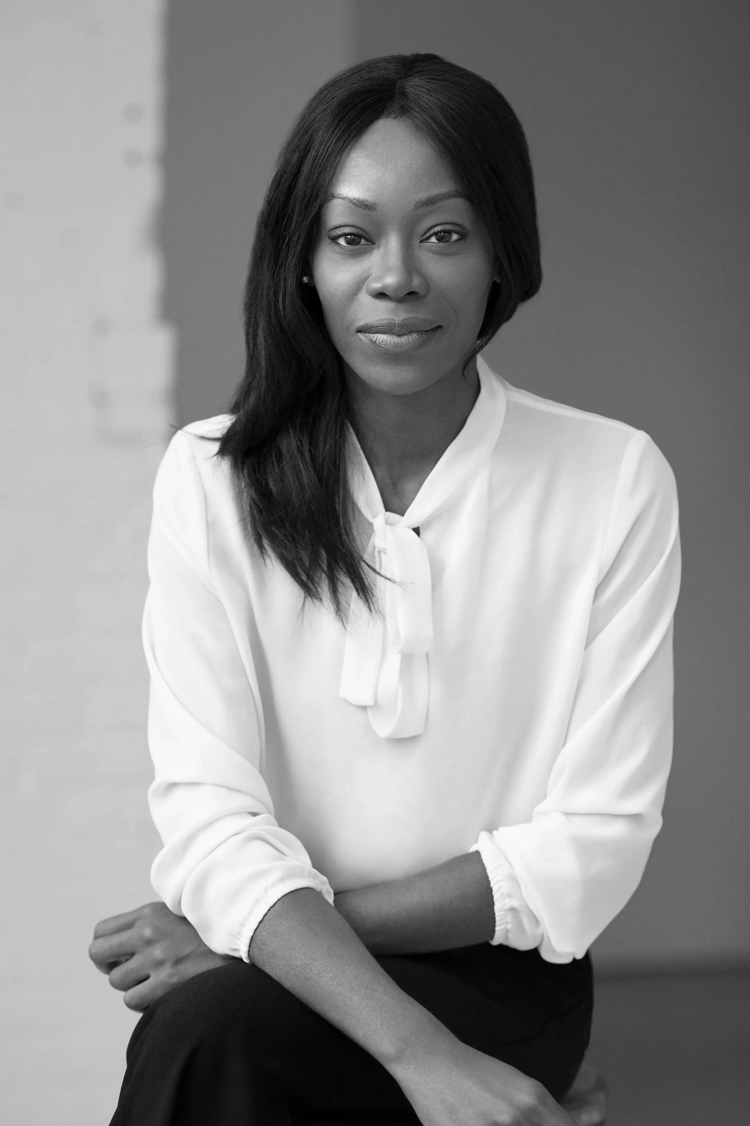 Black and white portrait of a confident woman with shoulder-length hair, wearing a white blouse with a bow at the collar, sitting with arms crossed, looking directly at the camera.
