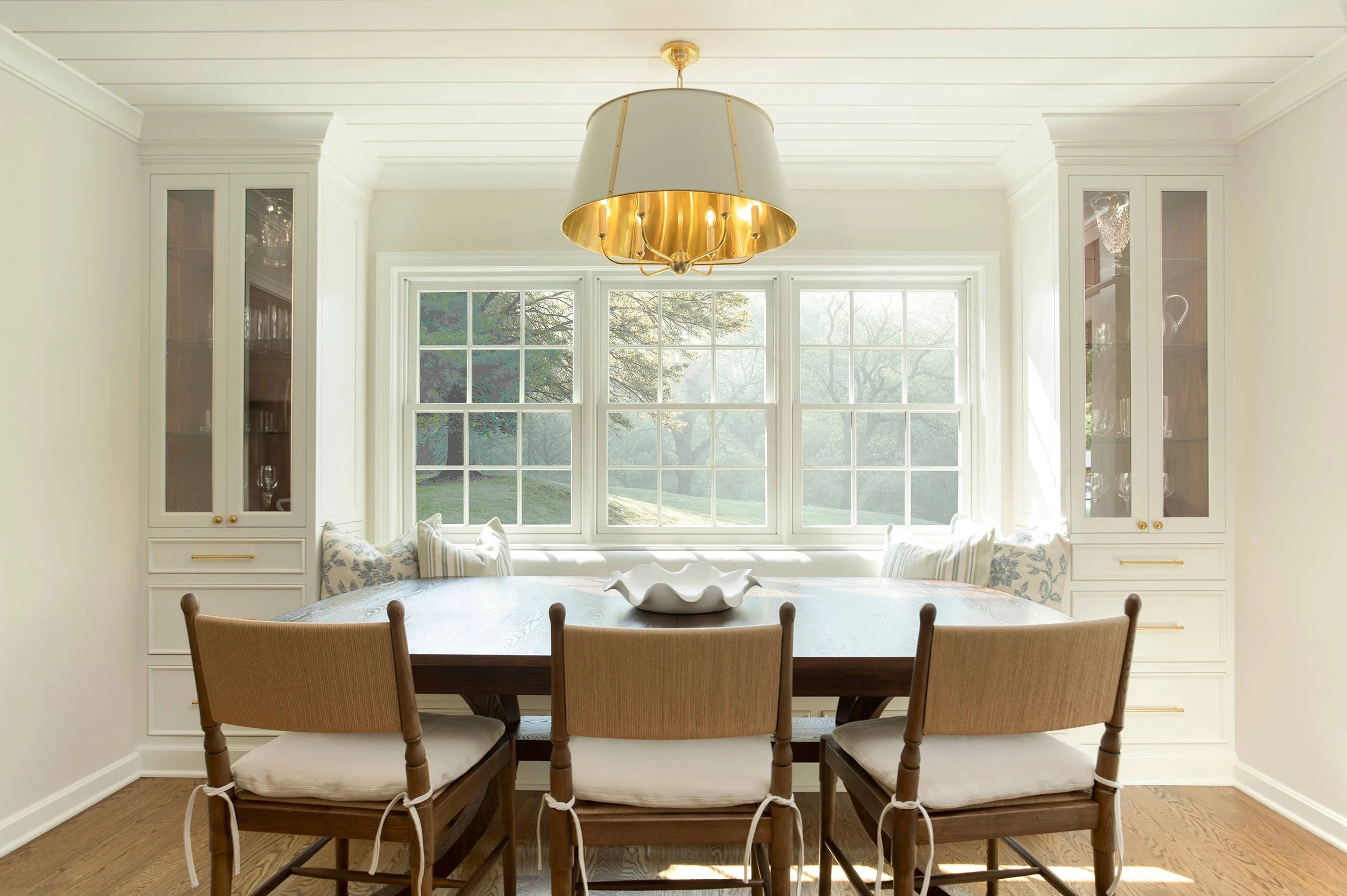 History Retold dining area in Shaker Heights, Ohio with a custom built-in banquette, white cabinetry, wood dining table, and brass lighting.