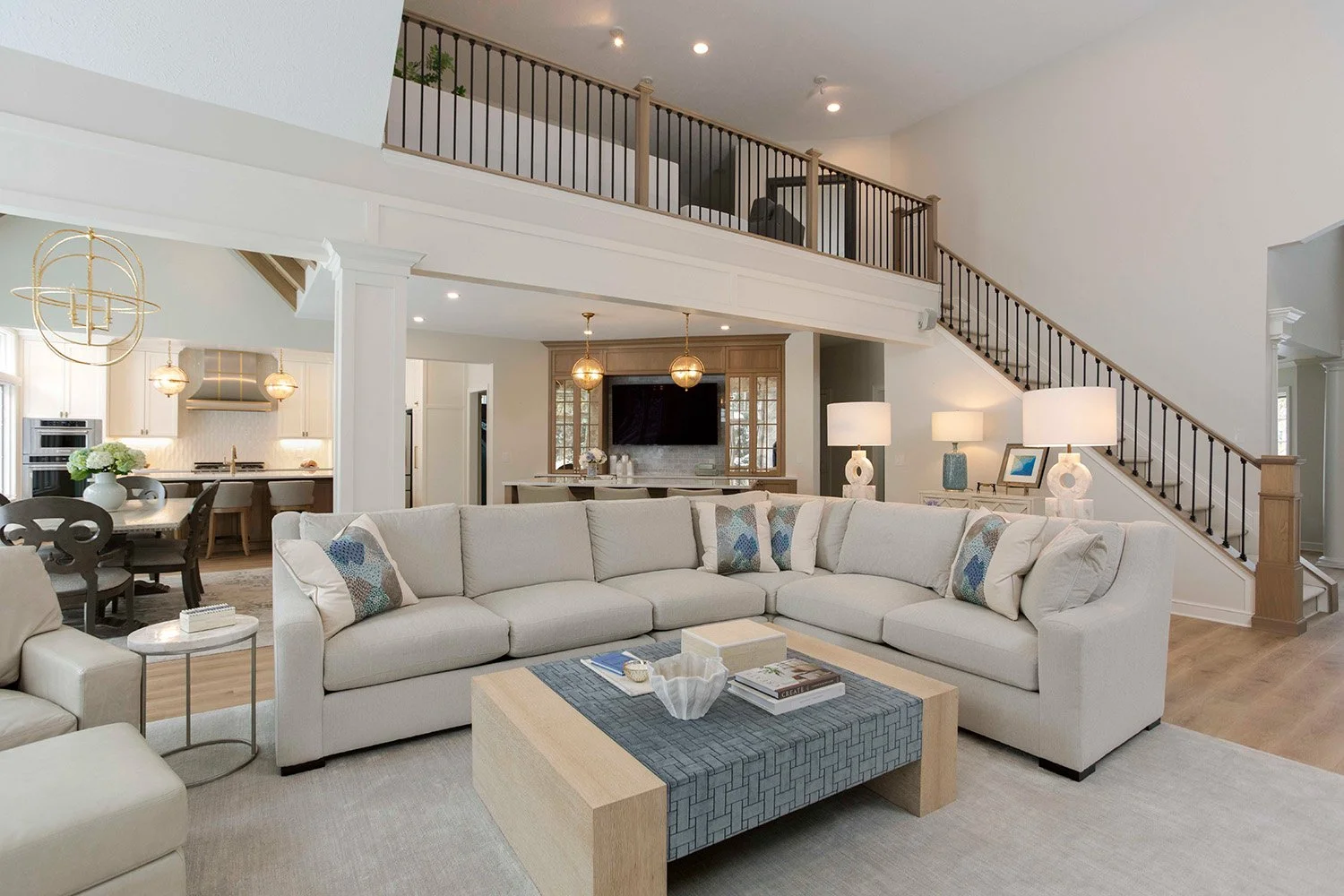 Expansive open-concept living room with a neutral sectional sofa, tufted ottoman, grand staircase, and large windows flooding the space with natural light | Ducy Design