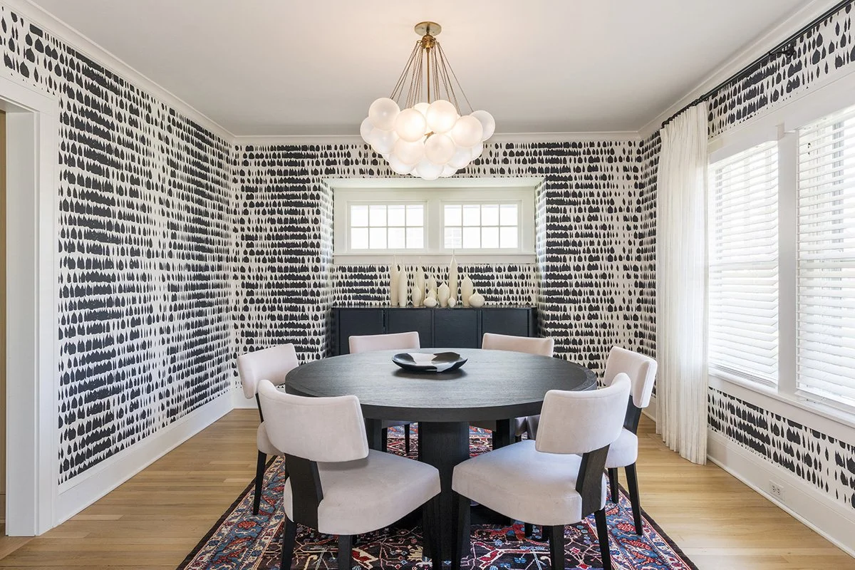 Dining room with bold black and white graphic wallpaper, a round dark table, upholstered white chairs, a globe chandelier, and a layered patterned rug | Ducy Design