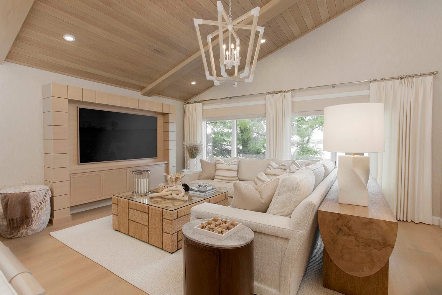 Open-concept lakehouse living and kitchen area with warm wood cabinetry, a media wall, neutral upholstered seating, and a woven chandelier overhead | Ducy Design