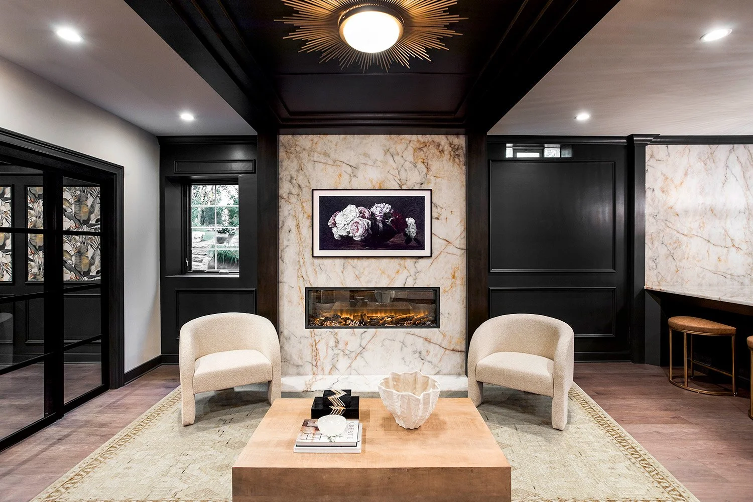 Intimate lounge seating area with two boucle accent chairs, a wood coffee table, a framed linear fireplace, and soft ambient lighting on dark walls | Ducy Design