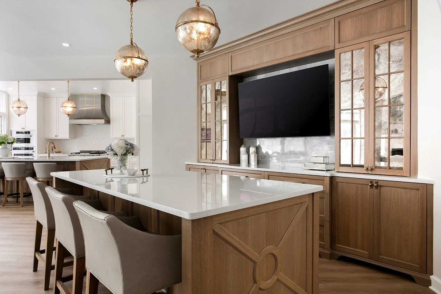 Open kitchen with warm oak cabinetry, a large quartz island with bar seating, antique mirrored accents, pendant lighting, and an integrated media wall | Ducy Design