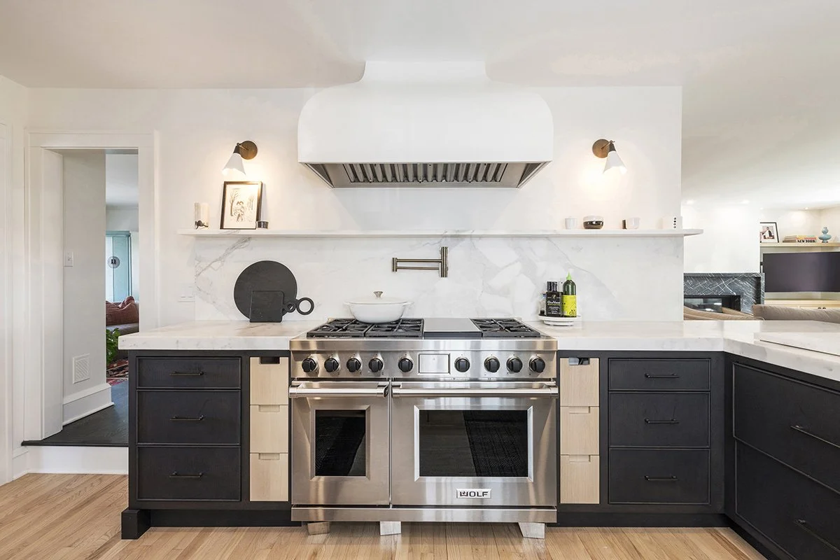 White kitchen with a professional-grade range, custom white hood, black lower cabinetry, open upper shelving, and a marble slab backsplash | Ducy Design