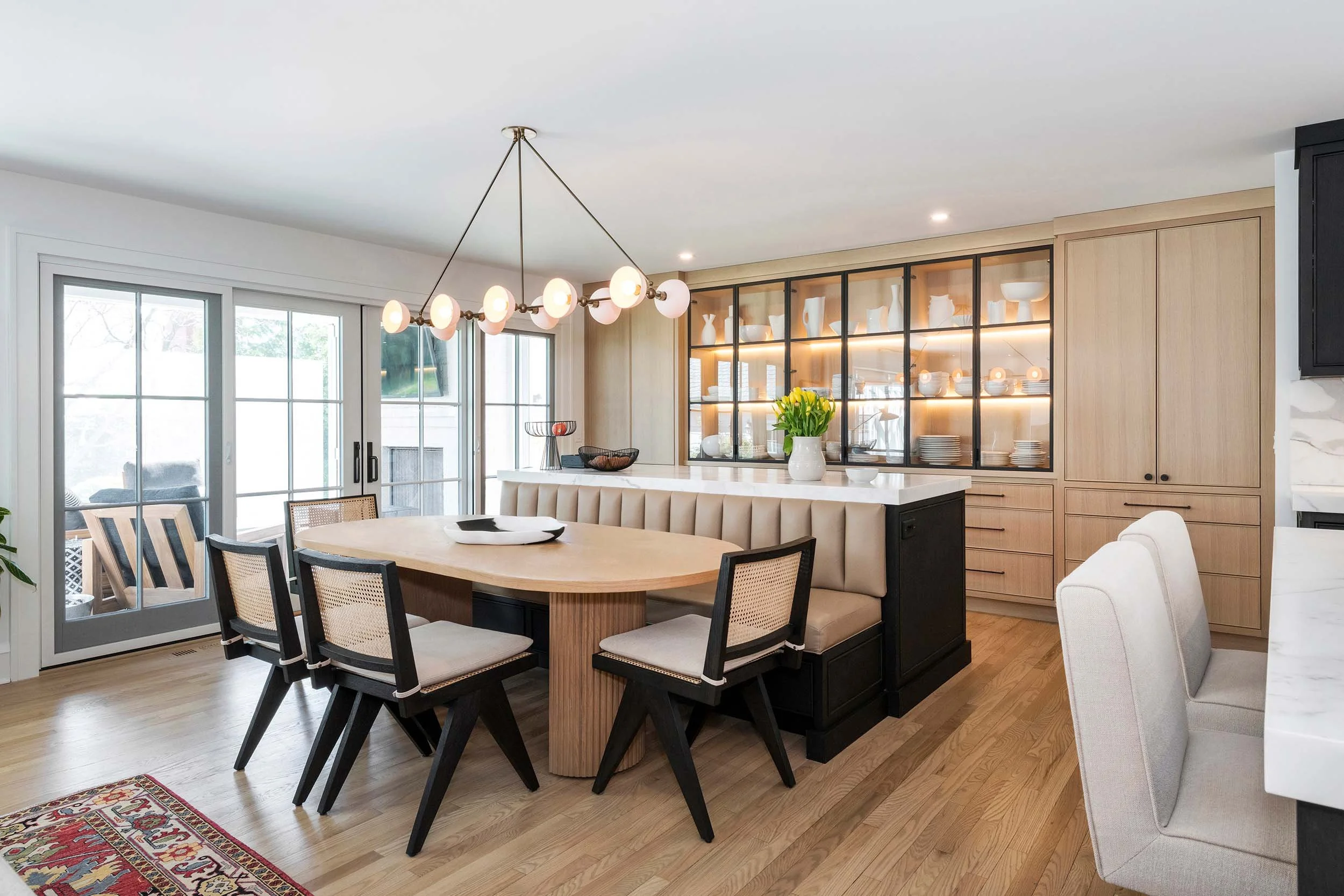 Modern Traditionalist dining area with sculptural banquette island, inset white oak cabinetry, iron-framed glass cabinets, and modern chandelier.