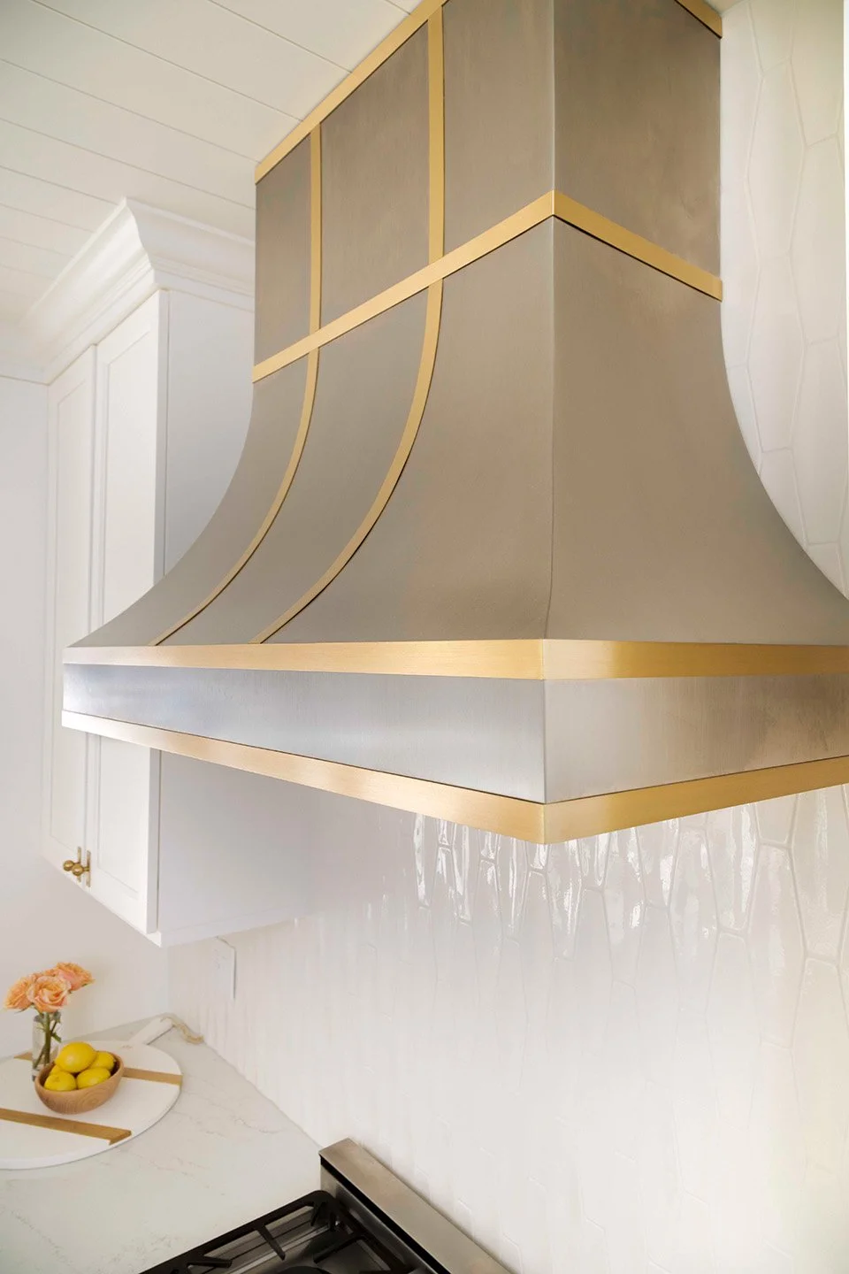Close-up of a custom plaster range hood with brushed gold geometric accent detail against white cabinetry and a professional range | Ducy Design