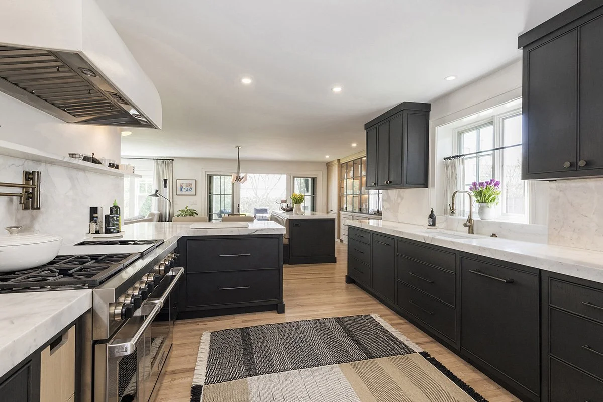 Wide-angle view of a dark kitchen with black cabinetry, a professional range, open shelving, and a connected living area with hardwood floors | Ducy Design