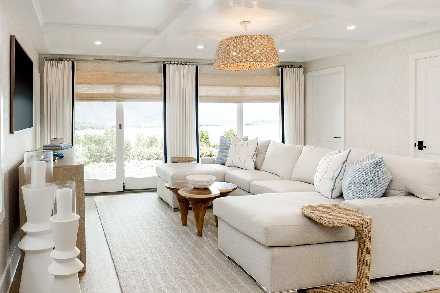 Bright coastal living room with a large white sectional, woven pendant light, natural Roman shades, and light blue accents in a lakehouse project by Ducy Design