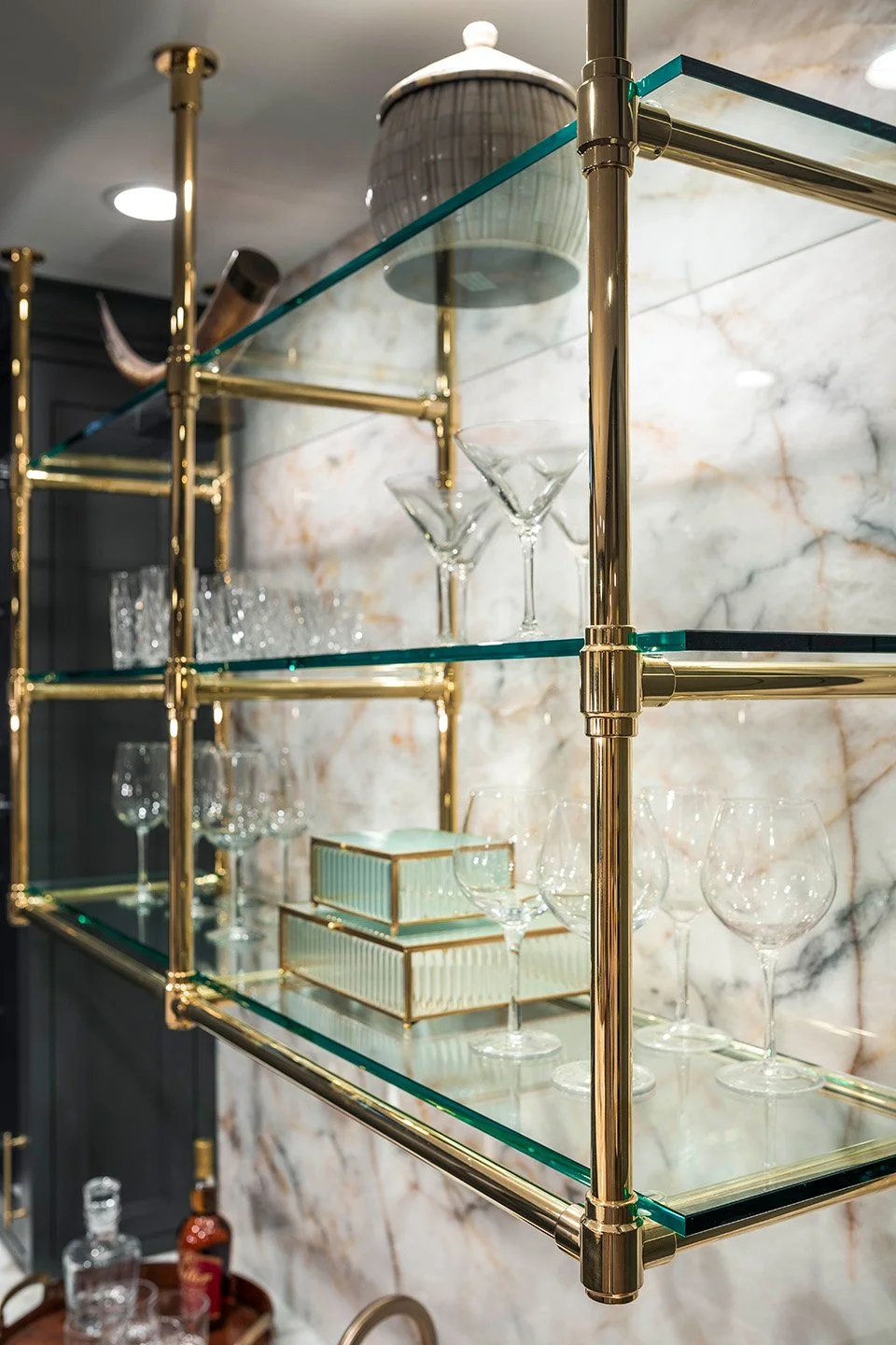 Full view of a custom angled brass and glass shelving unit styled with glassware and bottles against dark walls in the Luxe Lounge | Ducy Design