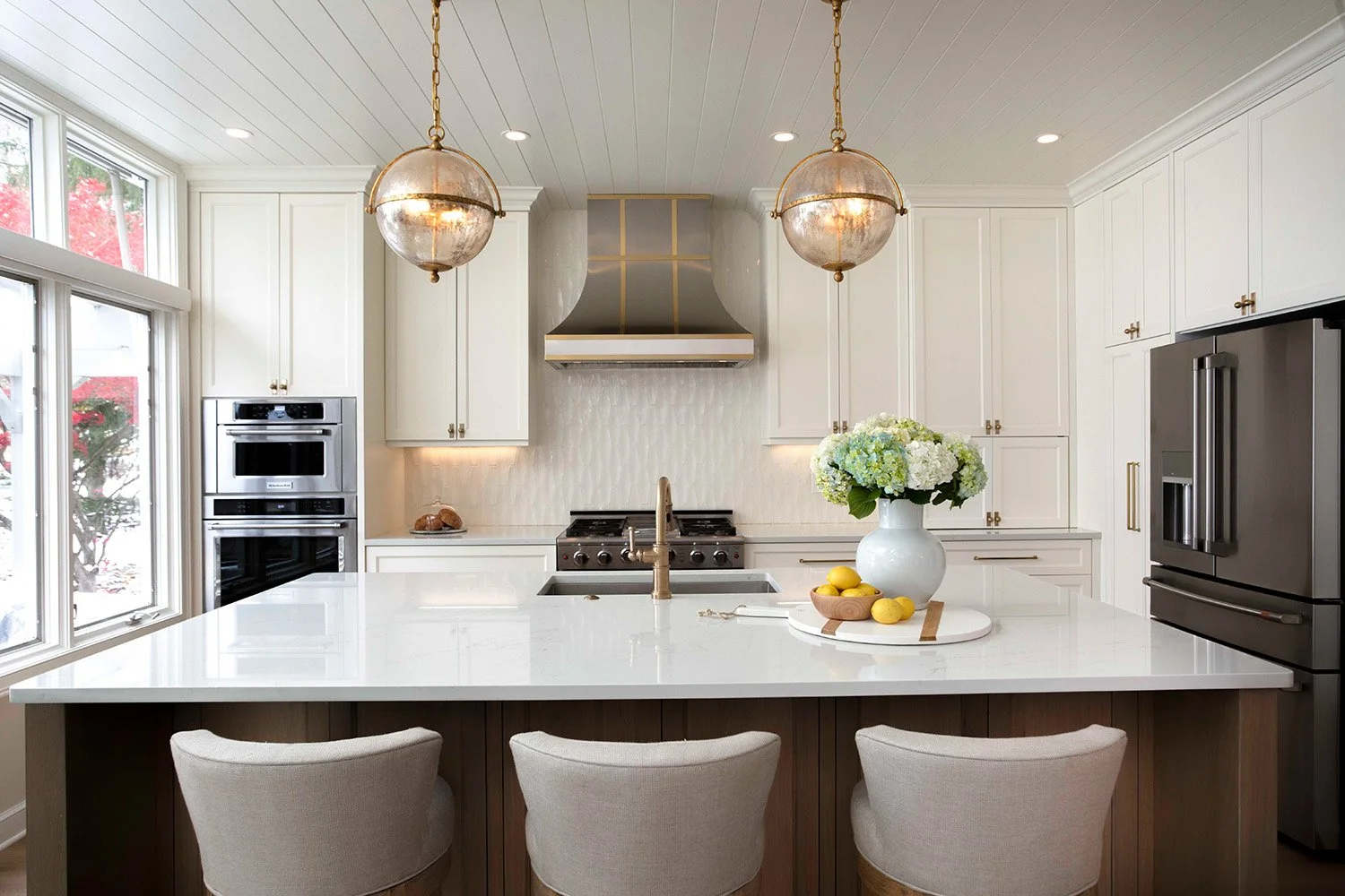 Styled kitchen island with white floral arrangement, upholstered bar stools, warm wood cabinetry, and pendant lighting in an open entertaining space | Ducy Design