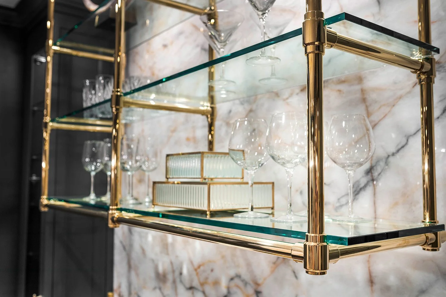 Close-up of a custom gold and glass bar shelving unit with angled brass frame, glass shelves, and wine glass storage against a dark backdrop | Ducy Design