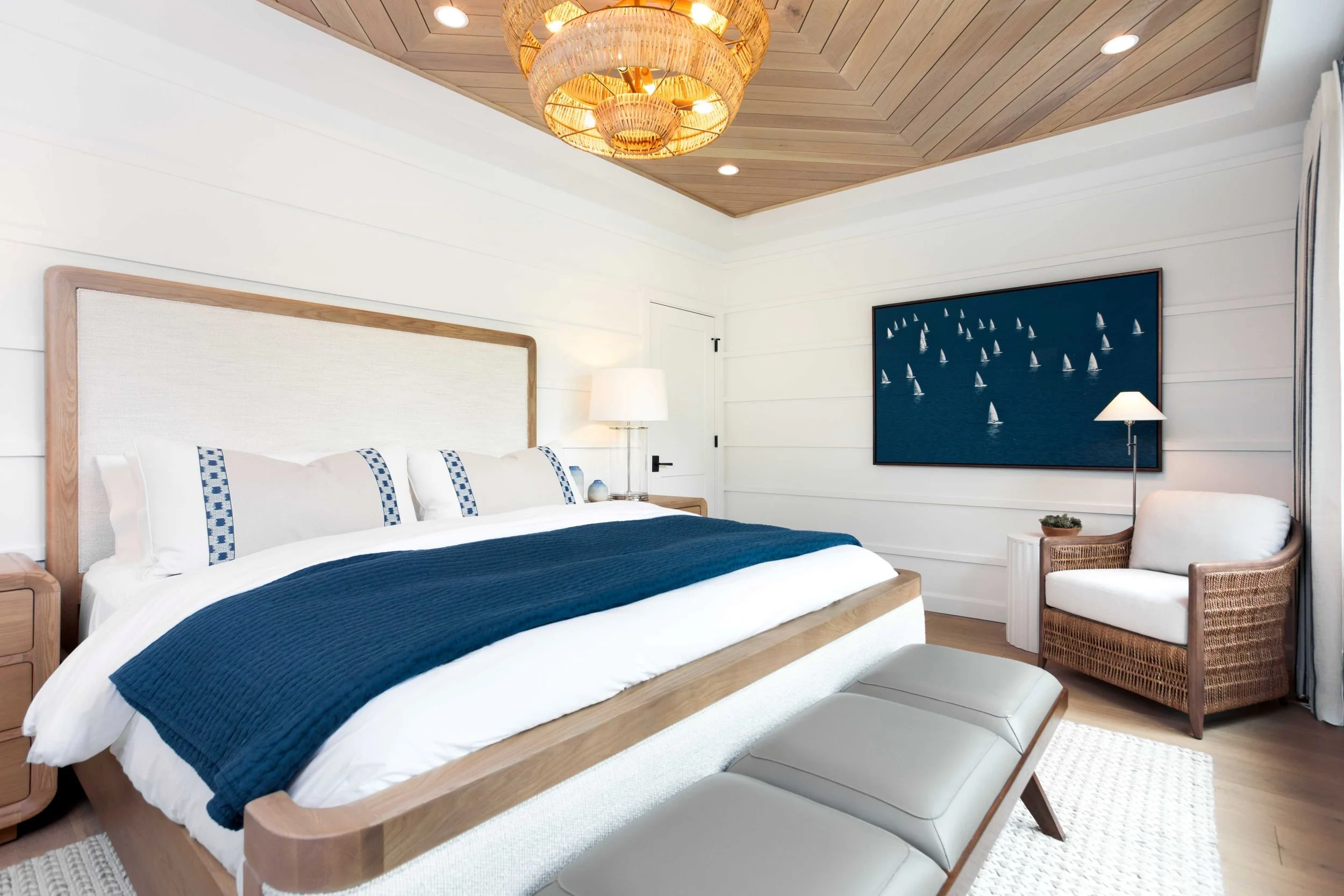 A primary bedroom with a herringbone wood ceiling, upholstered wood frame bed, navy blanket, and a sailboat themed artwork above a rattan chair | Ducy Design