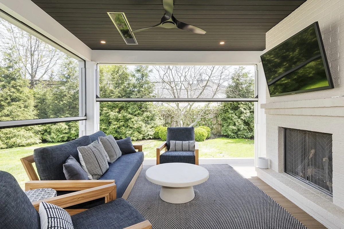 Screened porch with navy blue sofas, a white round coffee table, shiplap ceiling, and views of a lush green backyard through large windows | Ducy Design
