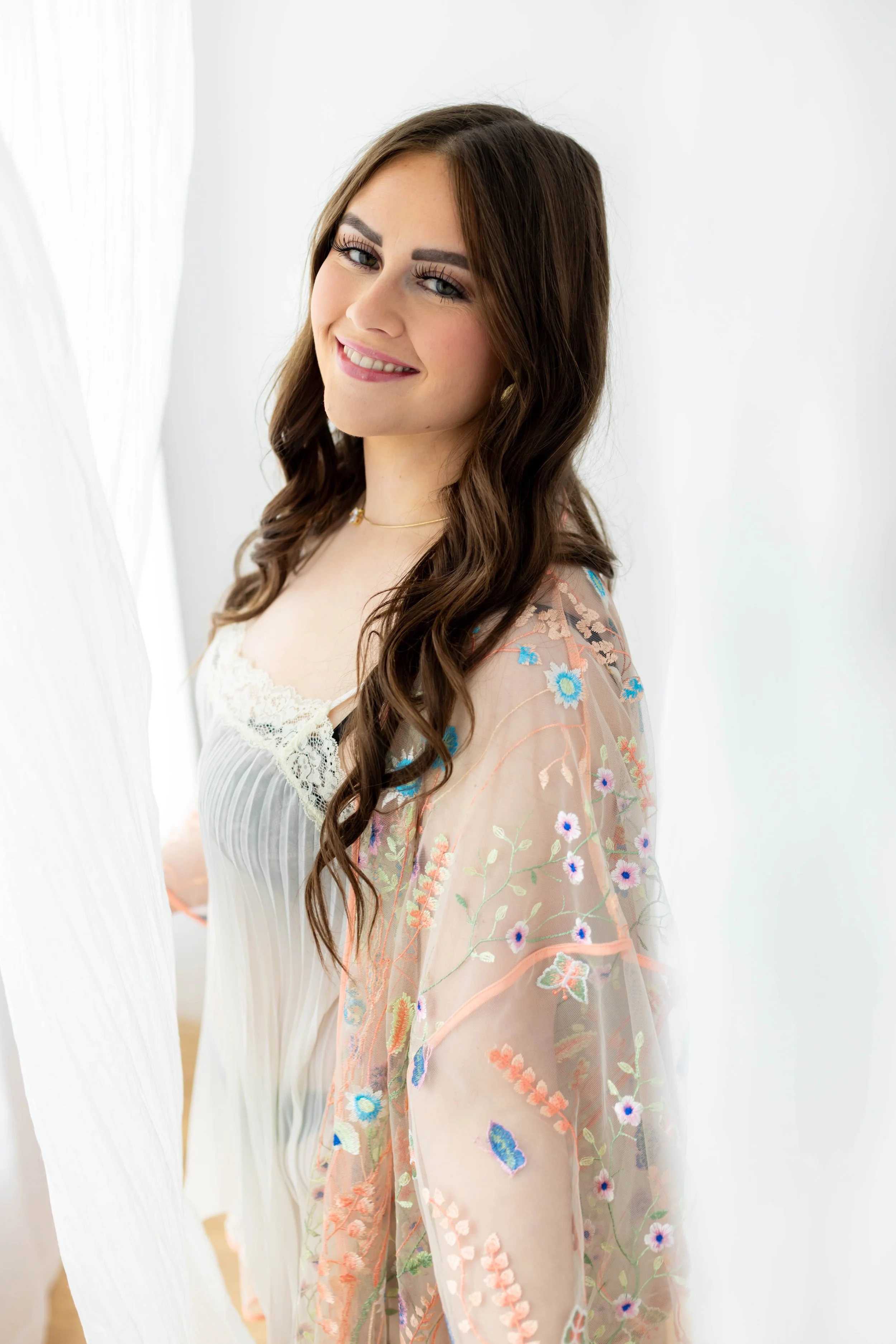 A woman looking into the camera as she stands in front of a window with white sheer curtains. She's wearing a brightly embrodered sheer robe over a pleated nightgown.