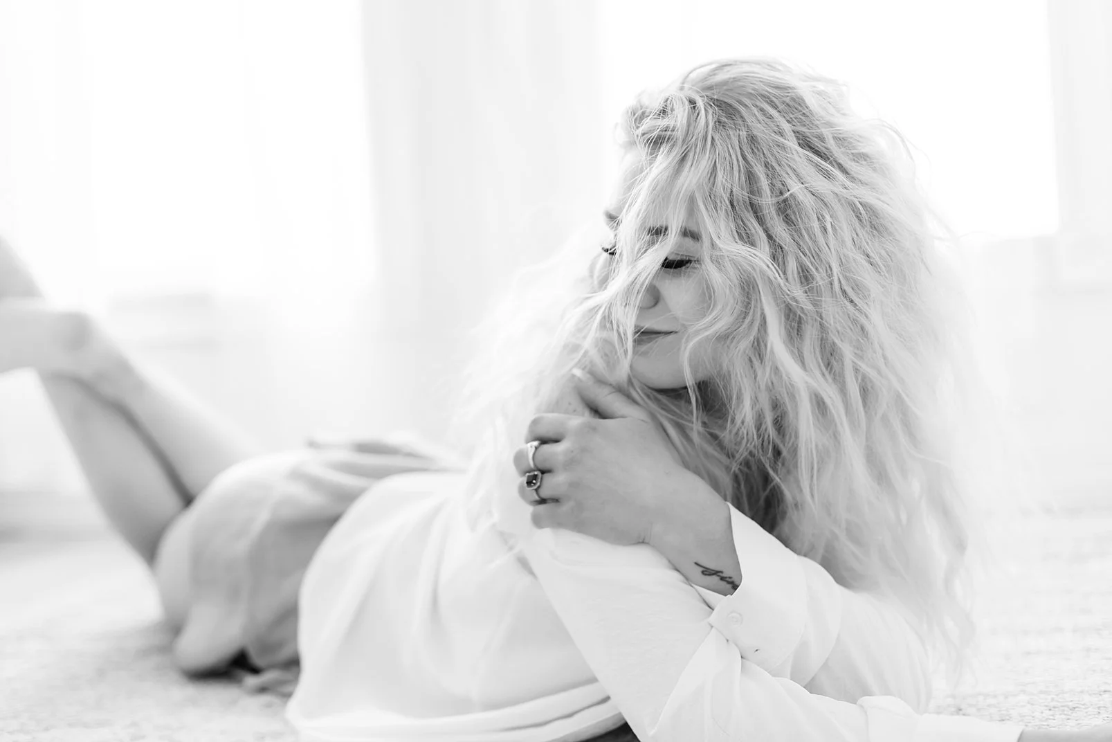 Woman in white blouse and a mini skirt laying on the floor, tummy down, supported by her elbows and giving herself a warm hug.