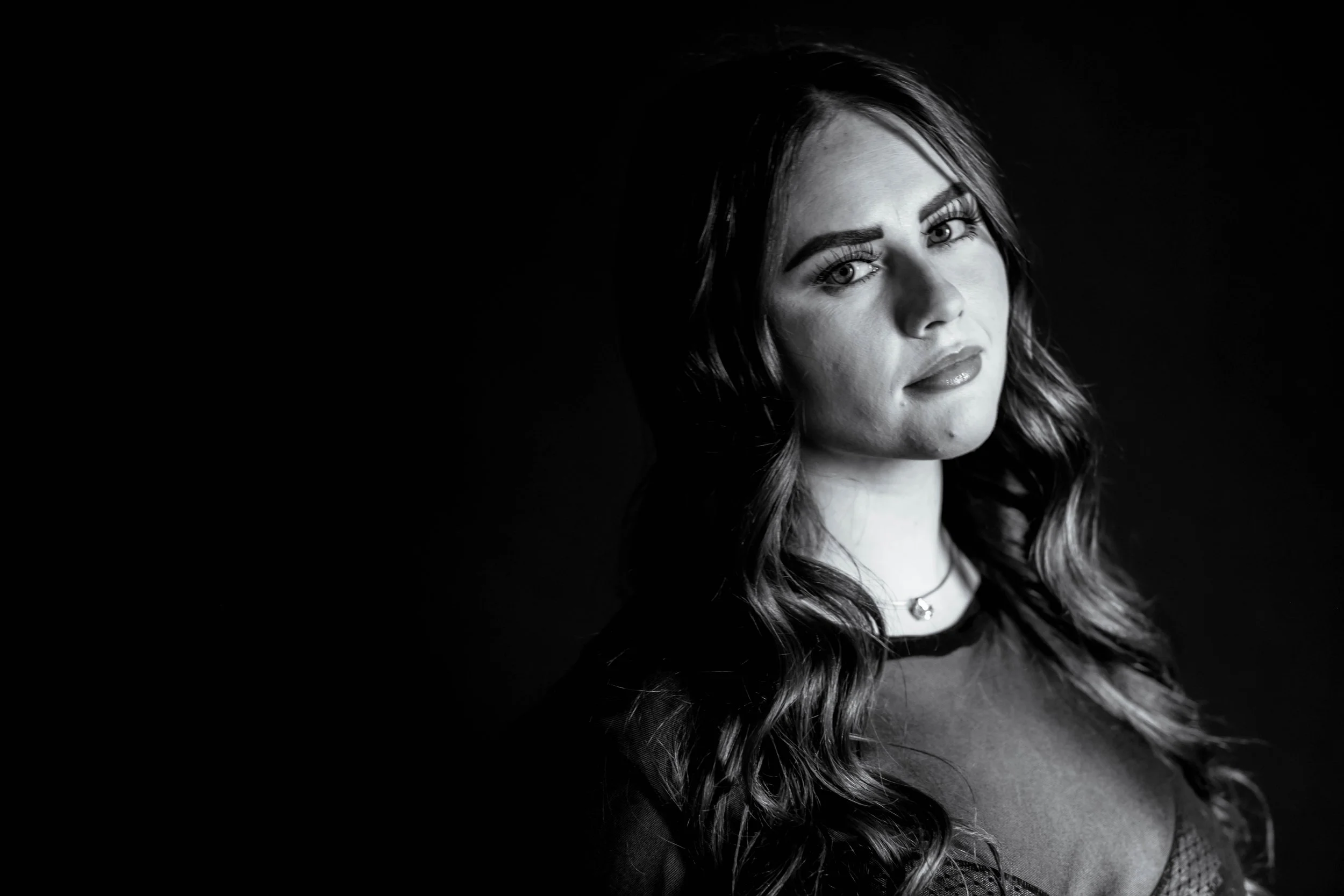 A beautiful brunet woman with flowing hair and black lingerie  looking into the camera with a slight smile. The background is dark, and she dramatically fades into it.