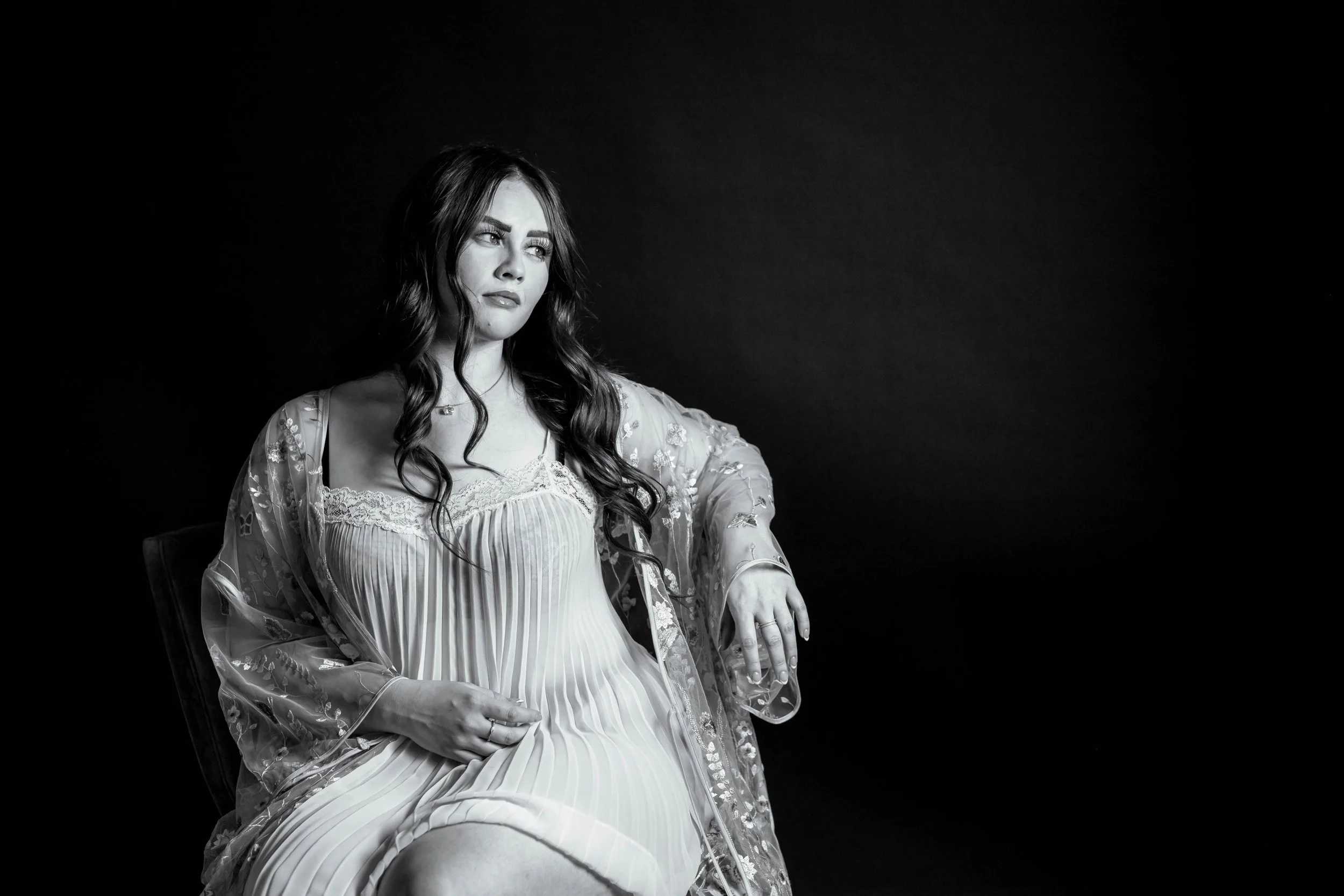 A beautiful woman wearing white lace sitting in a vintage chair with a black background. Her white lingerie pops in contrast in this black and white image.