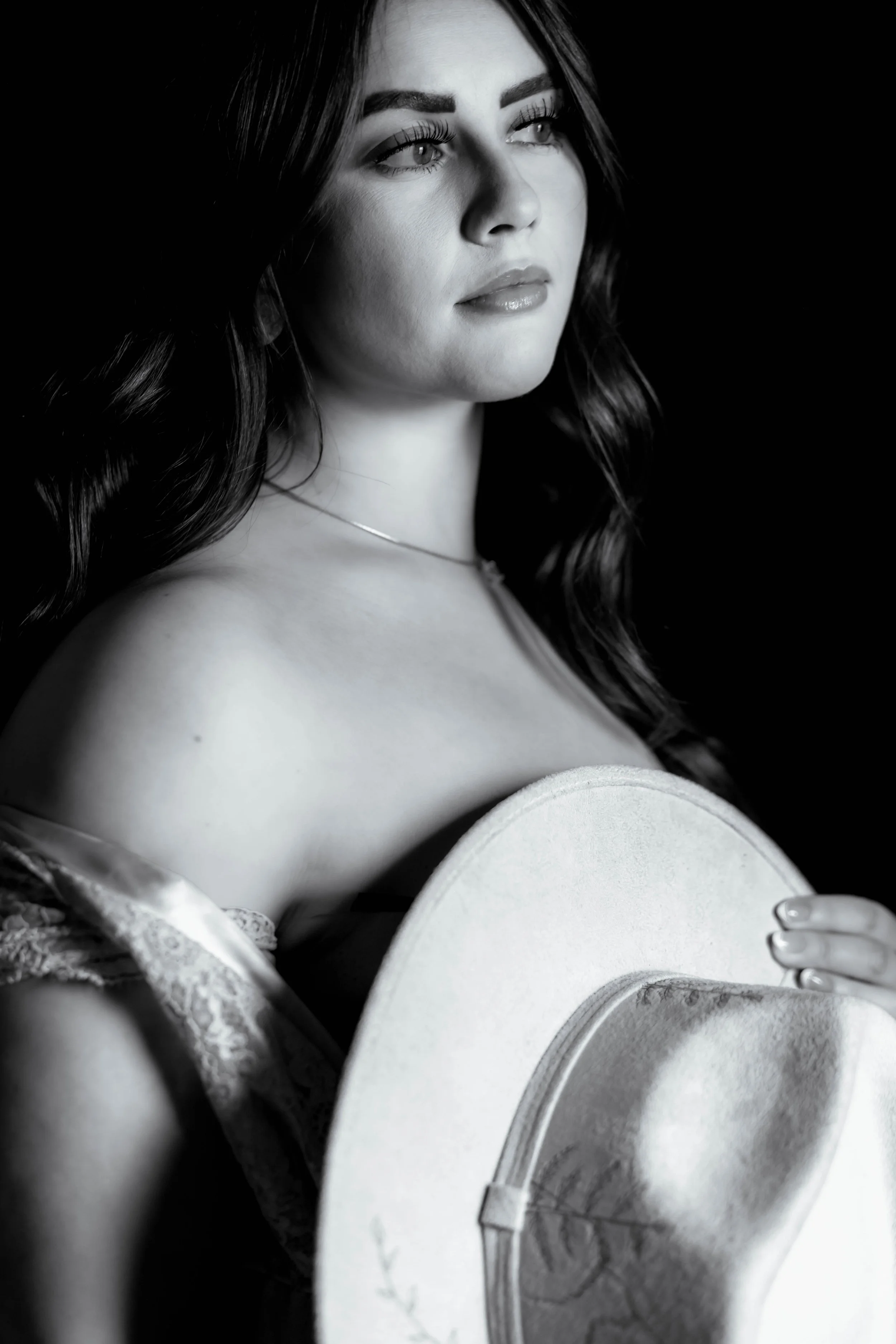 A black and white image of a dark haired woman with her robe falling off her shoulders and a delicately decorated hat covering her chest.