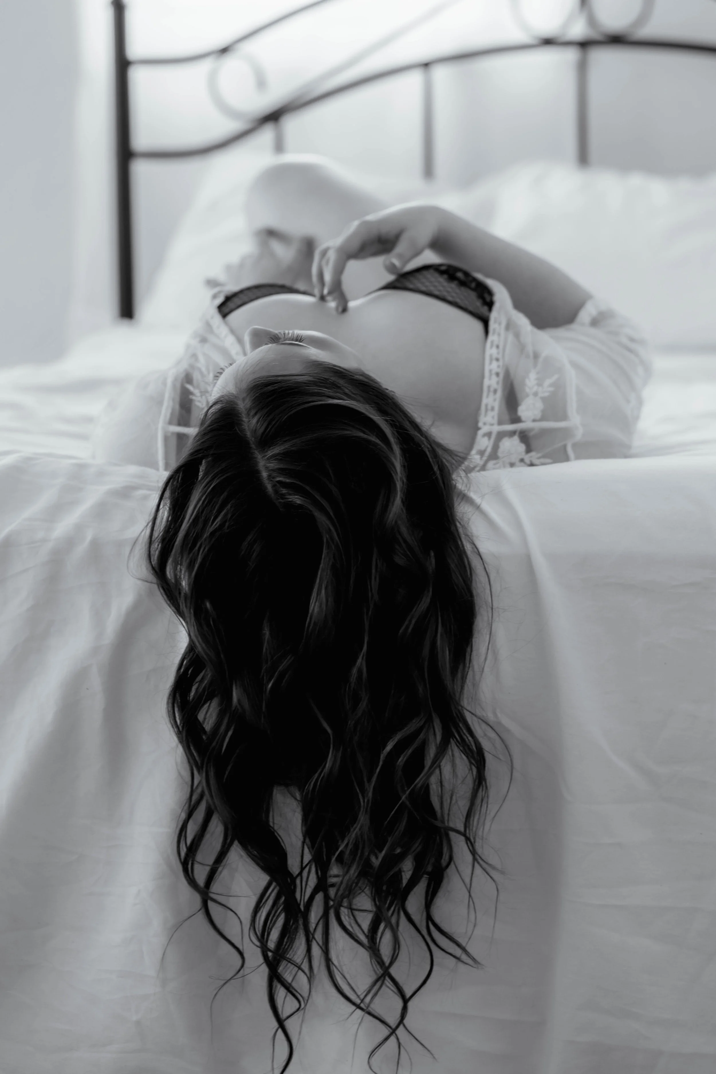 A woman laying on a bed with an iron headboard in the background. Her head is over the edge of the foot of the bed, hair flowing down. Her finger is trailing between her breasts, which are clothed in a black lace bra.