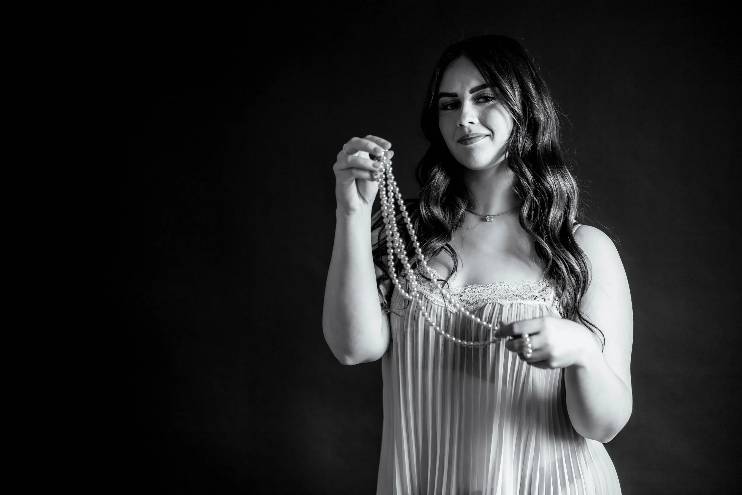 A woman in a white pleated short nighty, playing with a set of pearls in front of her and quietly smiling at the camera.