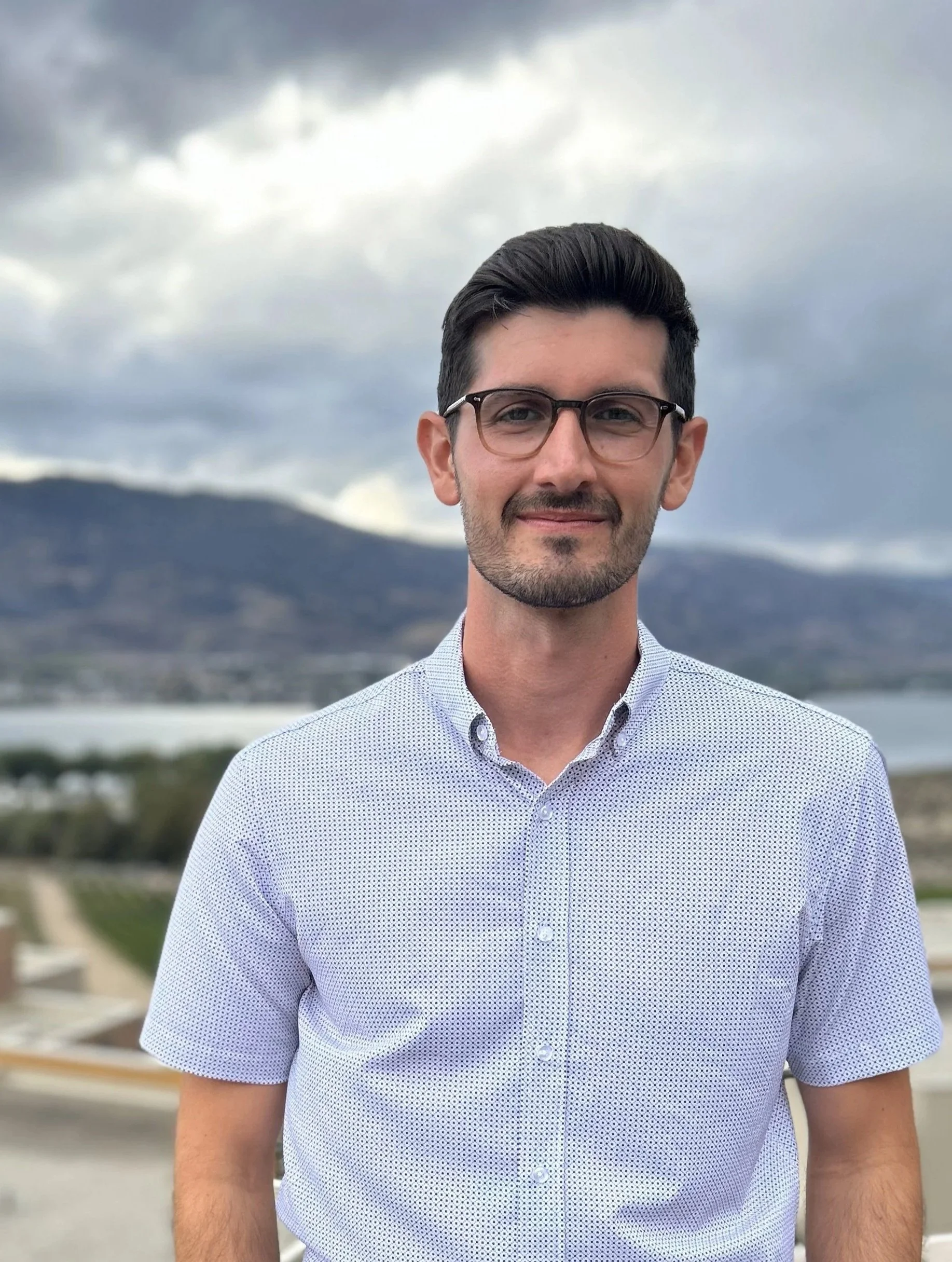 A man with short dark hair, glasses, and a beard, smiling outdoors with mountains, a lake, and cloudy sky in the background.