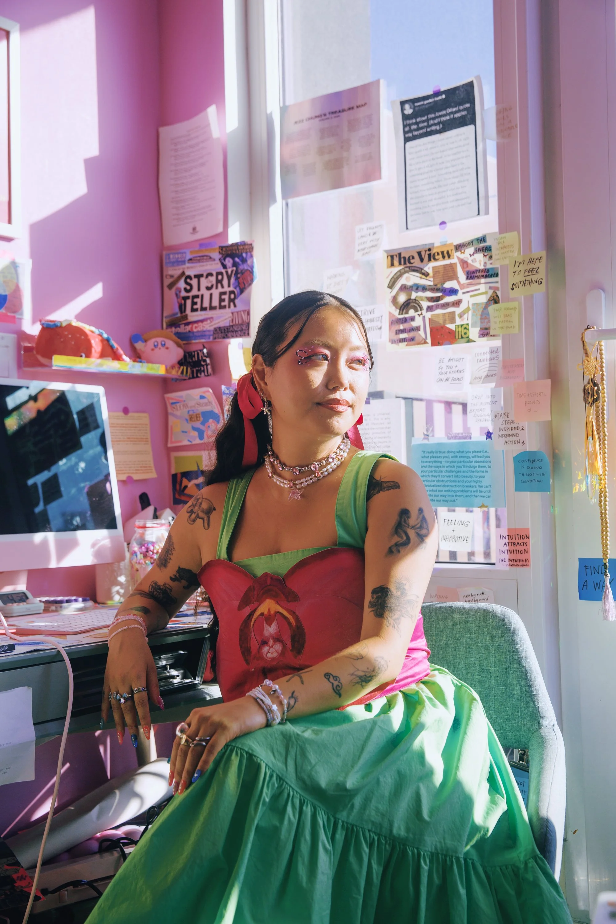 Photo of artist Jezz Chung at their former home office in Brooklyn, New York. They are surrounded by sticky notes with empowering affirmations and there is creative inspiration taped around the desk. The photo was taken by artist Mengwen Cao.