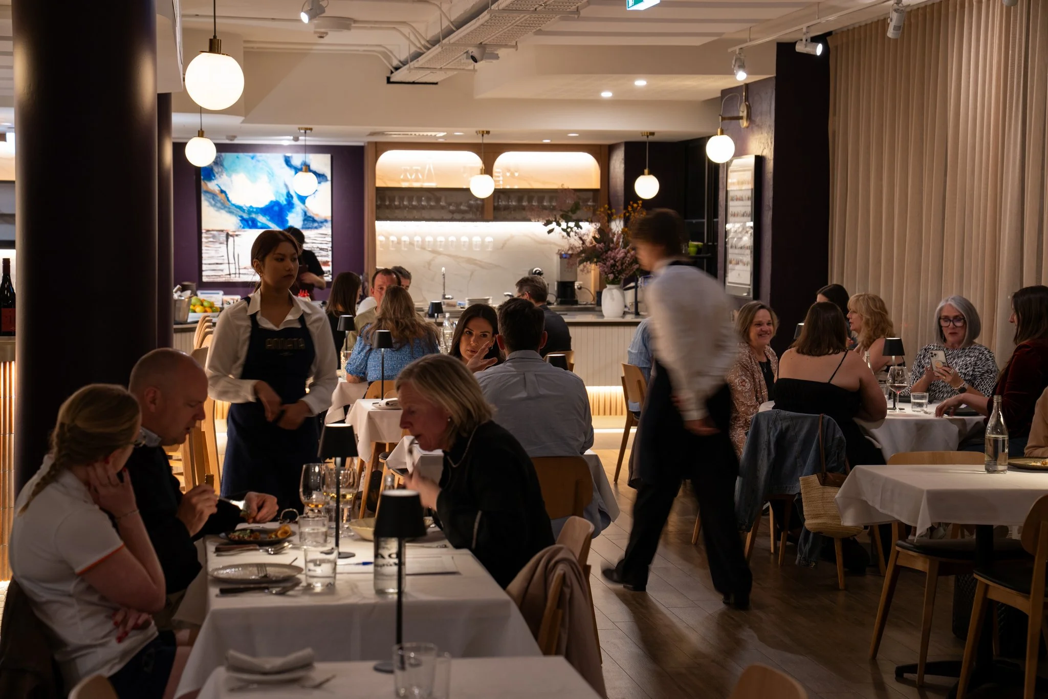 Guests dining in a modern upscale restaurant with waitstaff serving tables during a busy evening