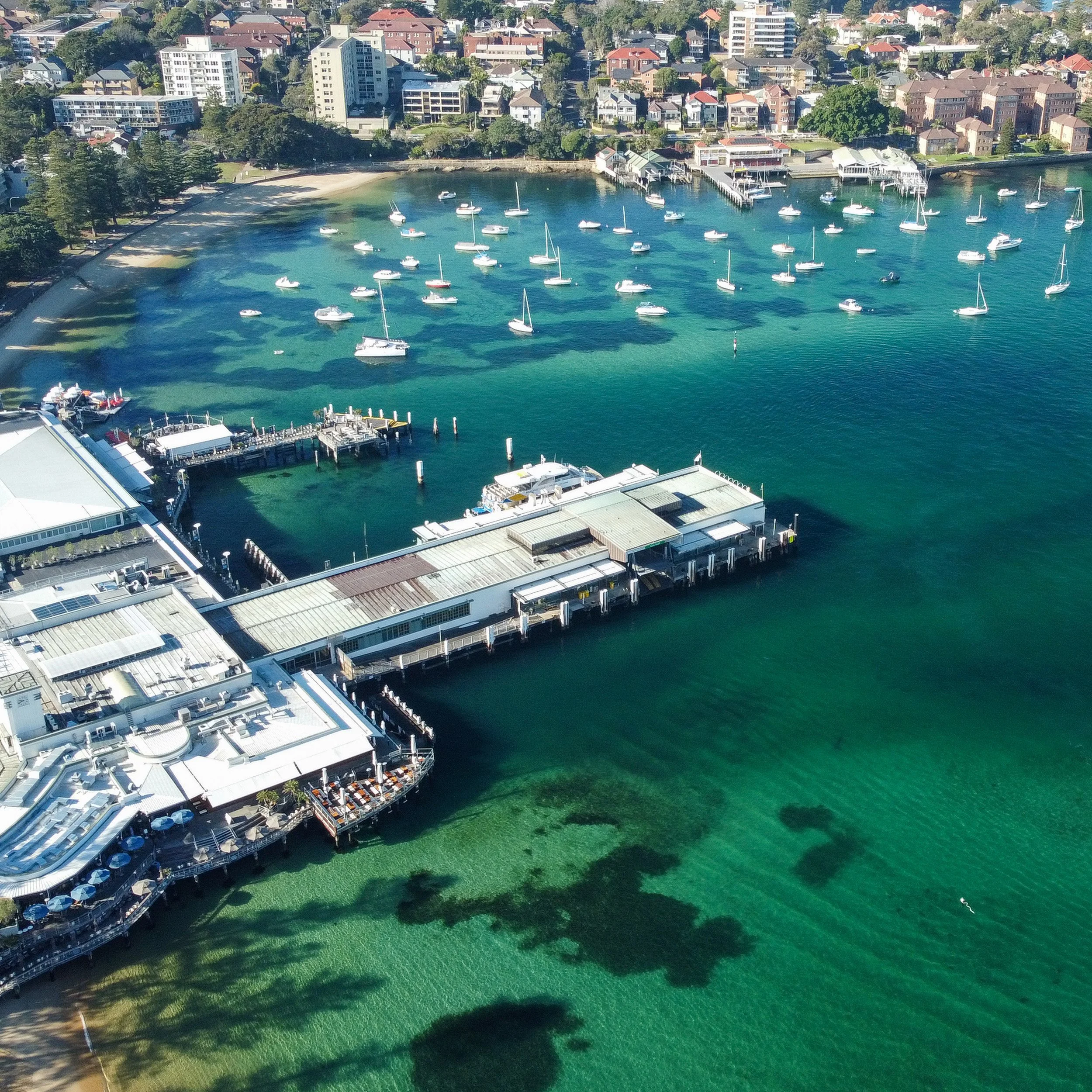 Aerial view of Manly Wharf with sailboats and yachts, adjacent to a city with residential buildings, trees, and a beach.