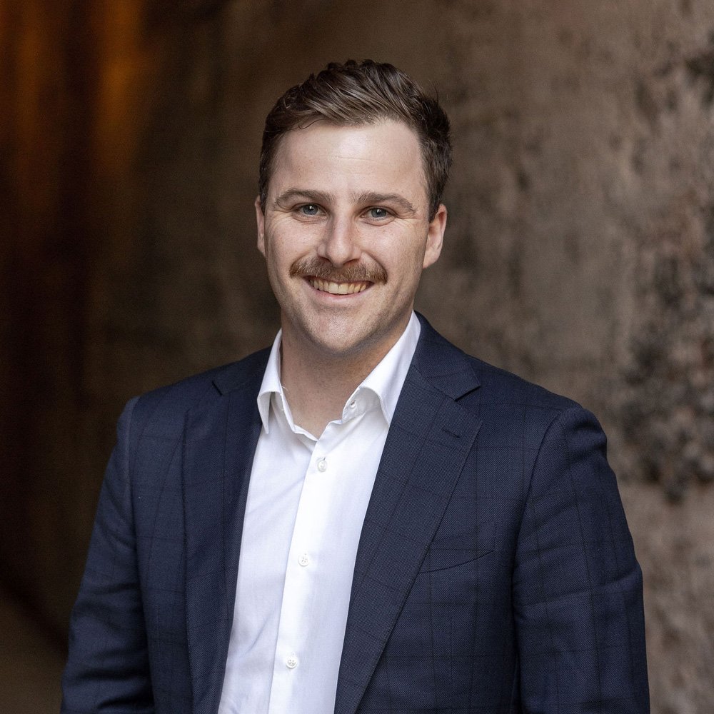A man with brown hair and a mustache smiling, dressed in a navy suit and white shirt, standing in front of a textured wall.