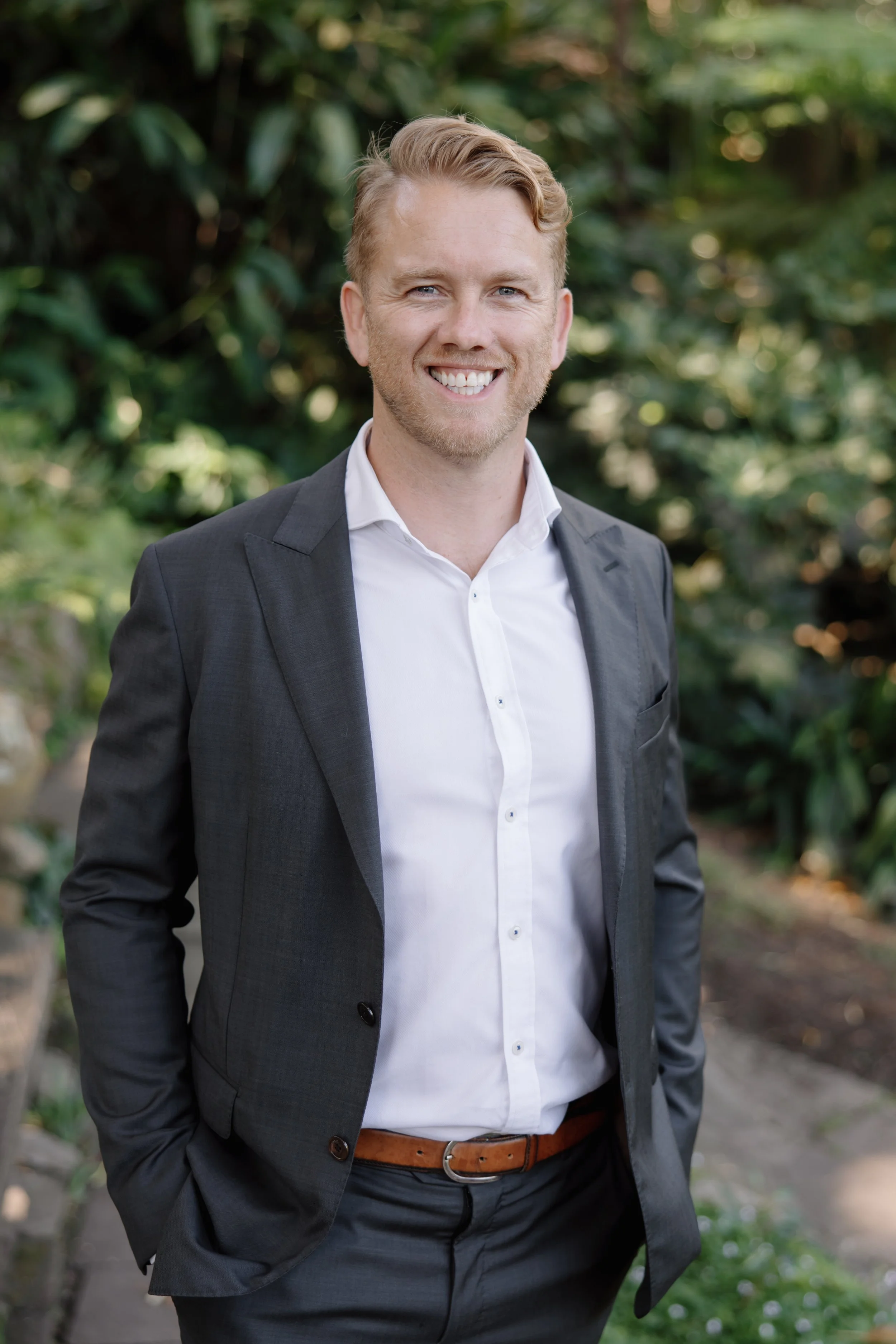 Smiling man in a suit standing outdoors with greenery in the background.