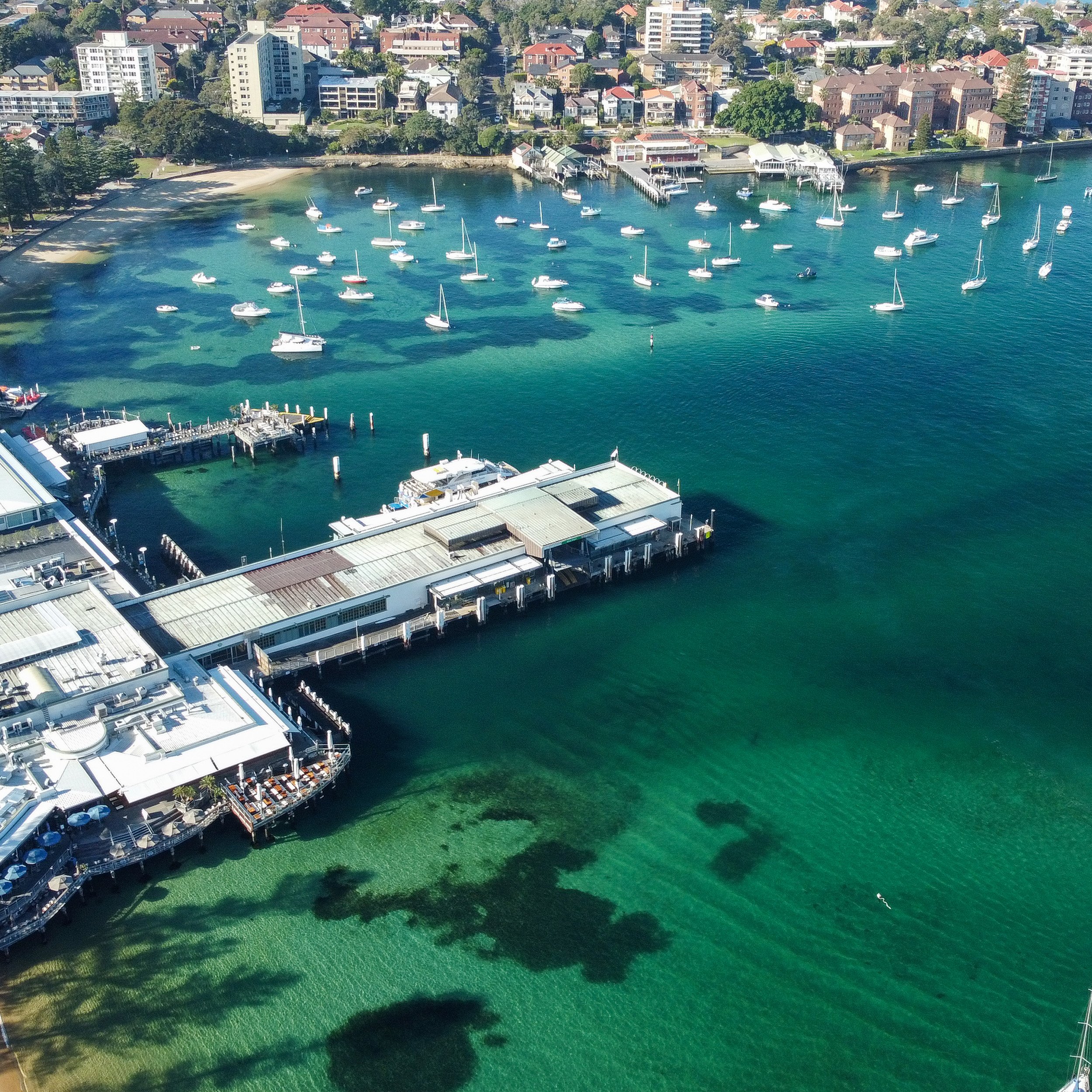 Aerial view of a marina with numerous sailboats and yachts docked in clear blue-green water, with a backdrop of residential houses and buildings on a hillside.