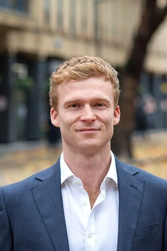 A man with light hair wearing a blue suit and white shirt, standing outdoors with blurred trees and buildings in the background.
