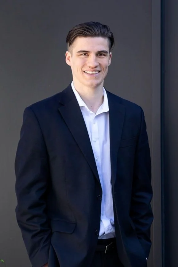 Young man in a navy suit and white shirt smiling, standing outdoors against a gray background.