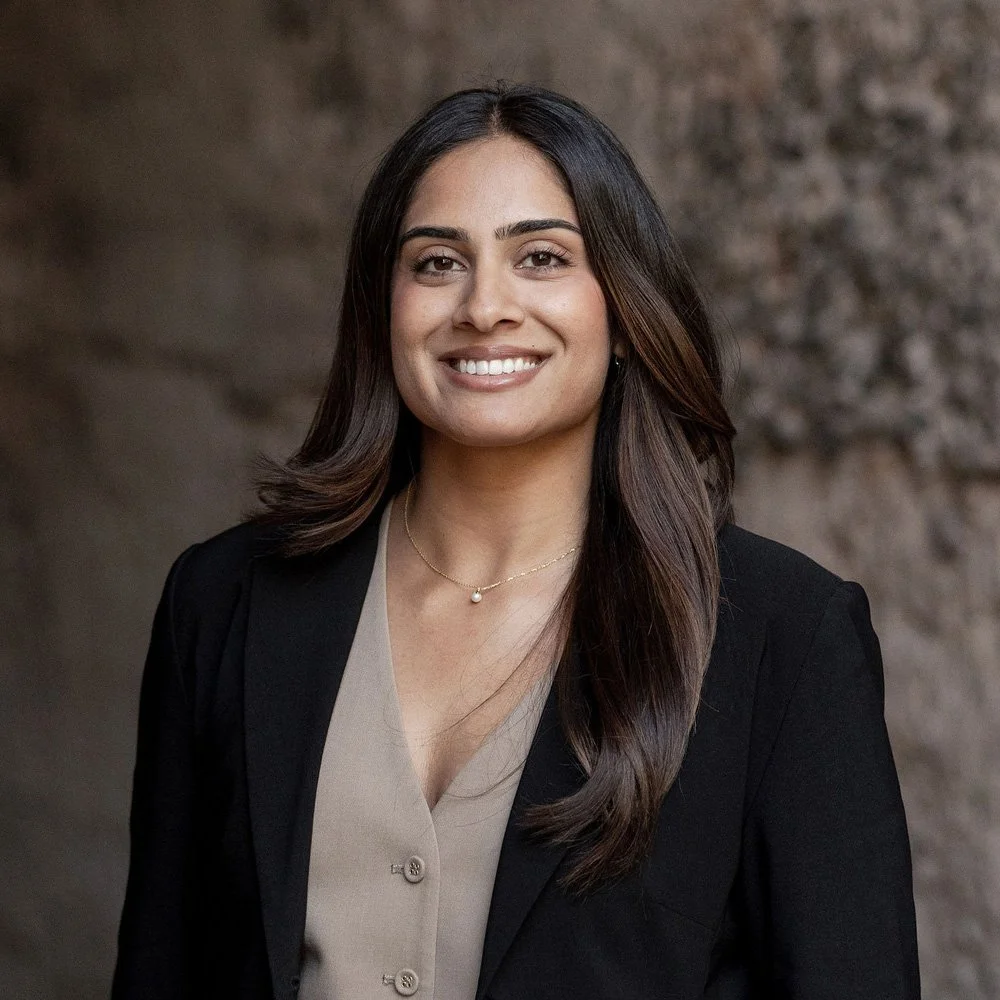 A woman with long dark hair wearing a black blazer and beige blouse, smiling at the camera. The background is blurred.