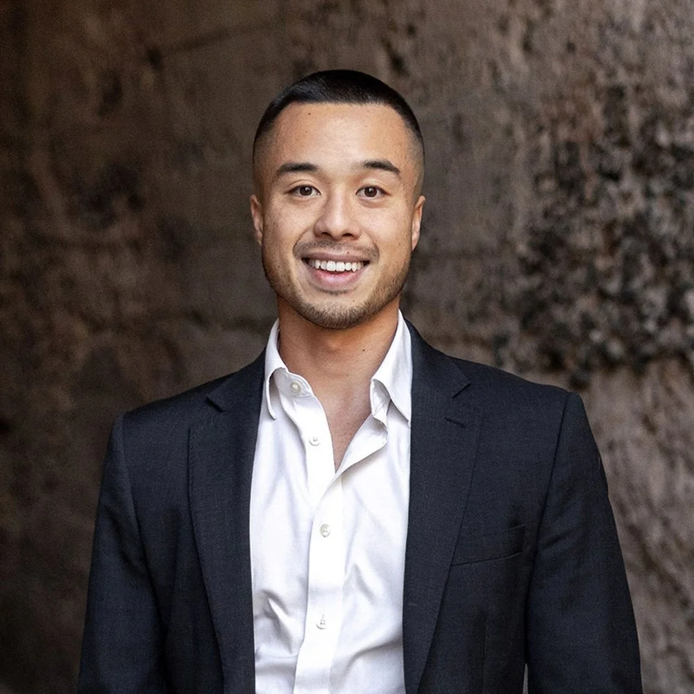 A man in a dark suit and white shirt smiling outdoors with greenery in the background.