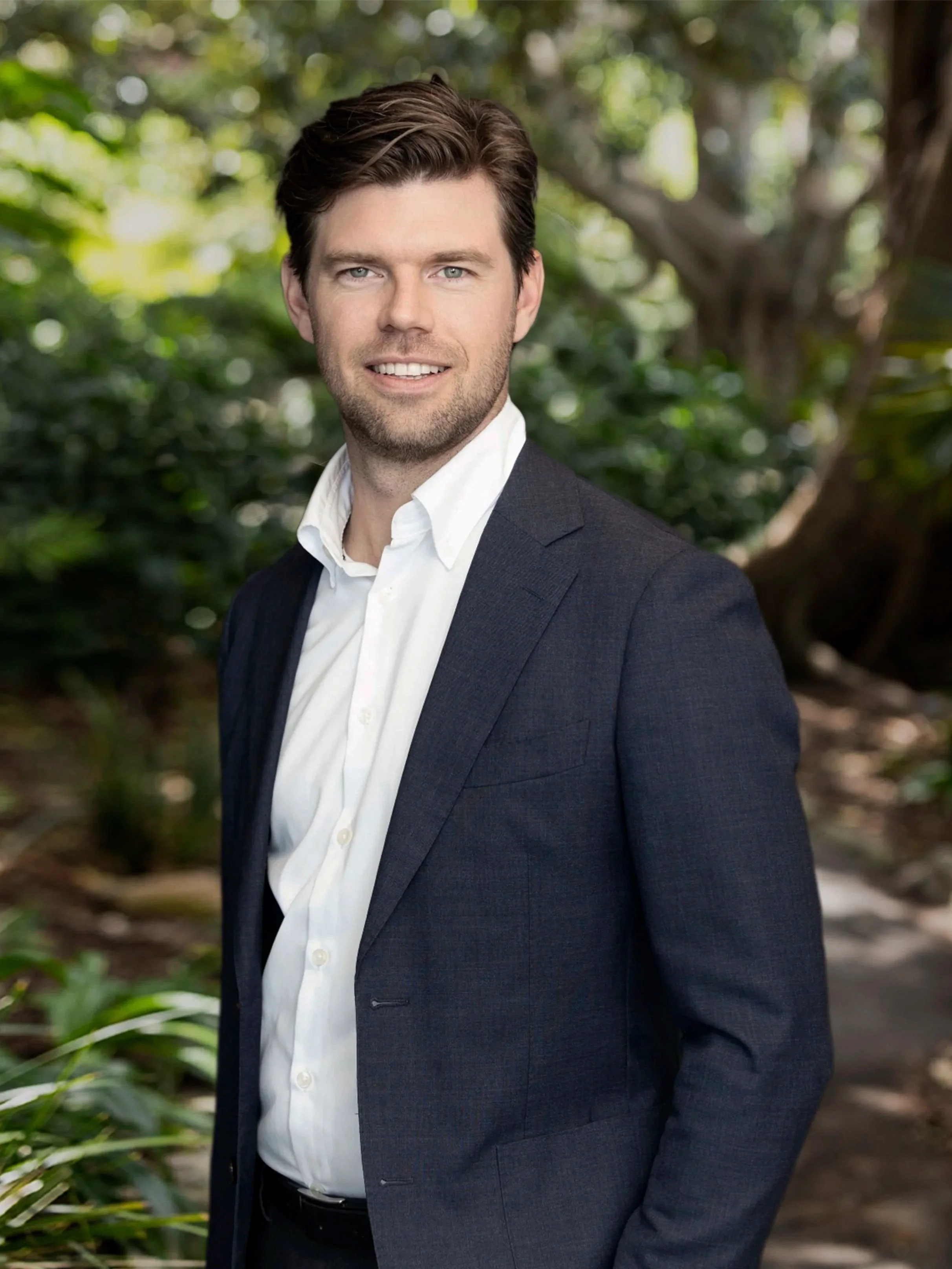 A young man with dark hair, wearing a navy blazer over a white shirt, smiling and standing against a dark wall.
