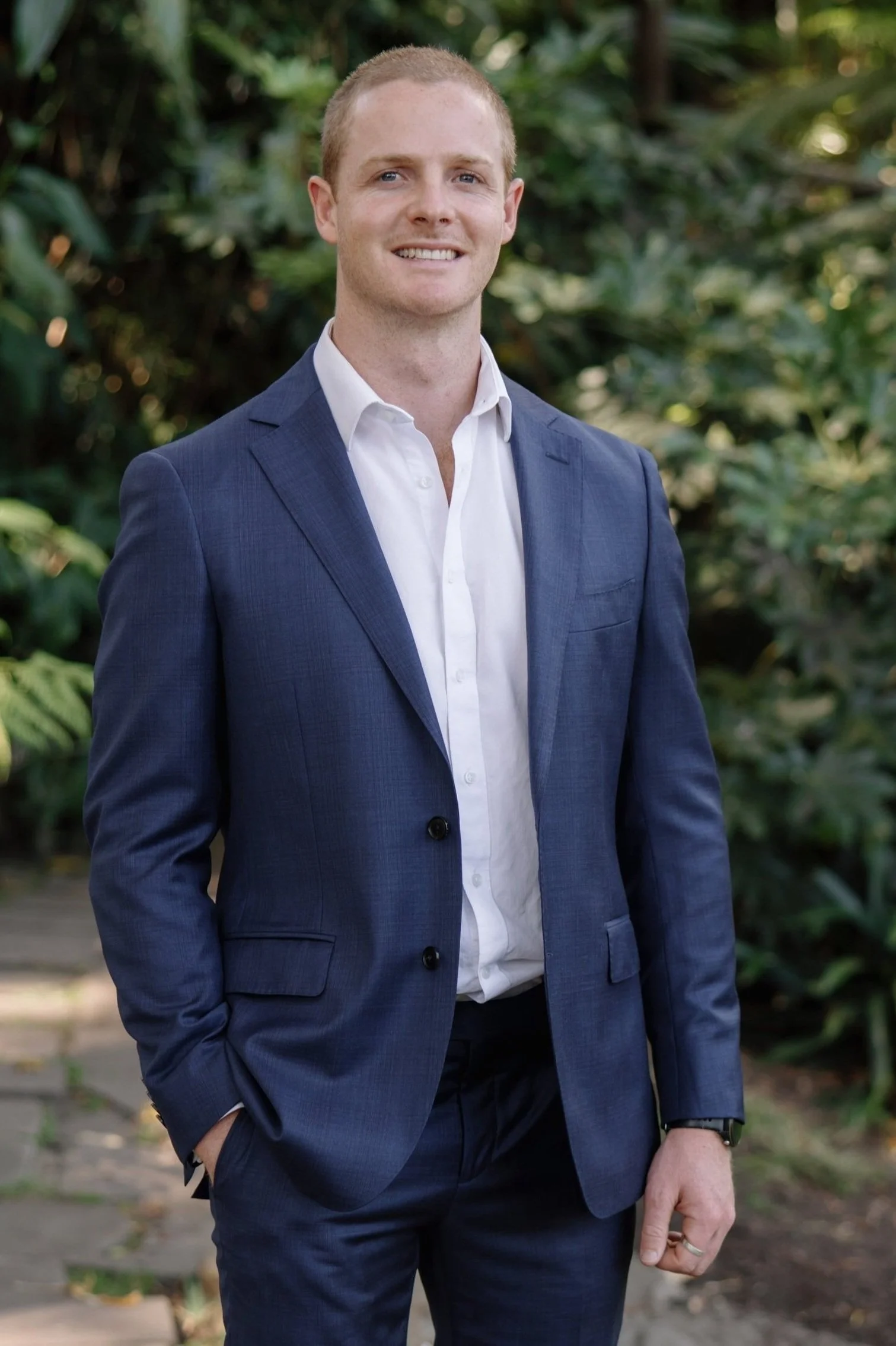 A man in a navy blue suit and white shirt standing outdoors with green foliage in the background.