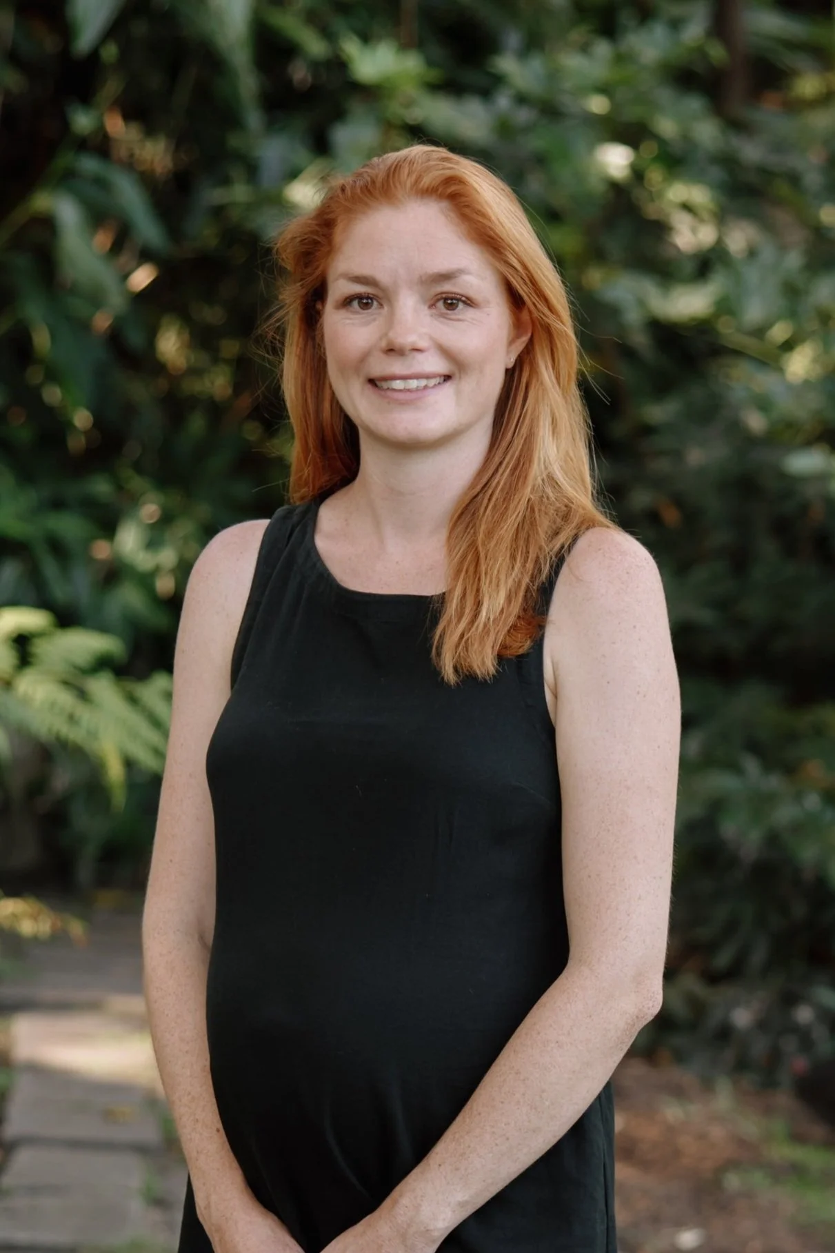 A woman with red hair wearing a black sleeveless dress, standing outdoors with greenery in the background, smiling at the camera.