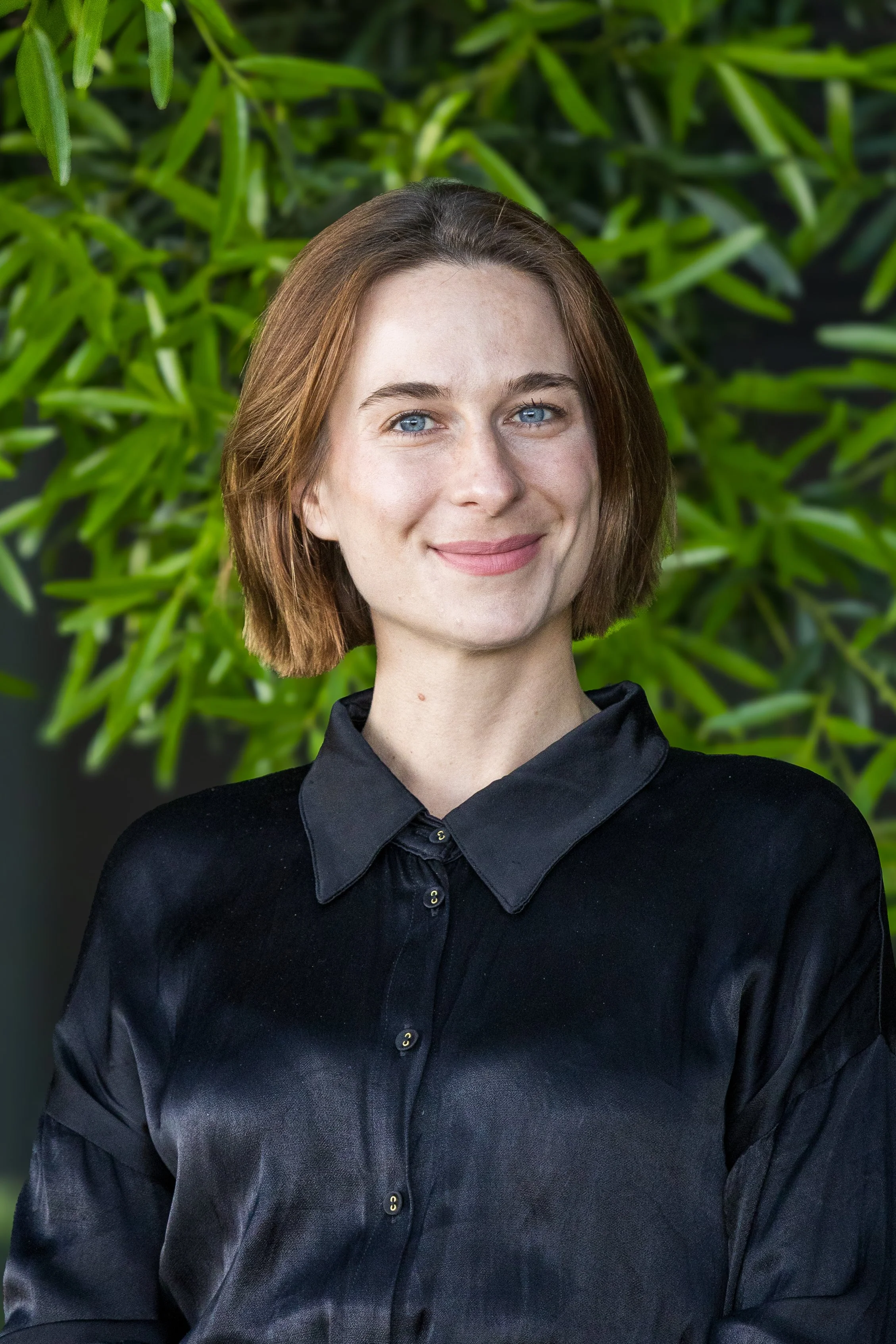 A woman with short brown hair and blue eyes, smiling, wearing a black satin button-up shirt, standing in front of green foliage.