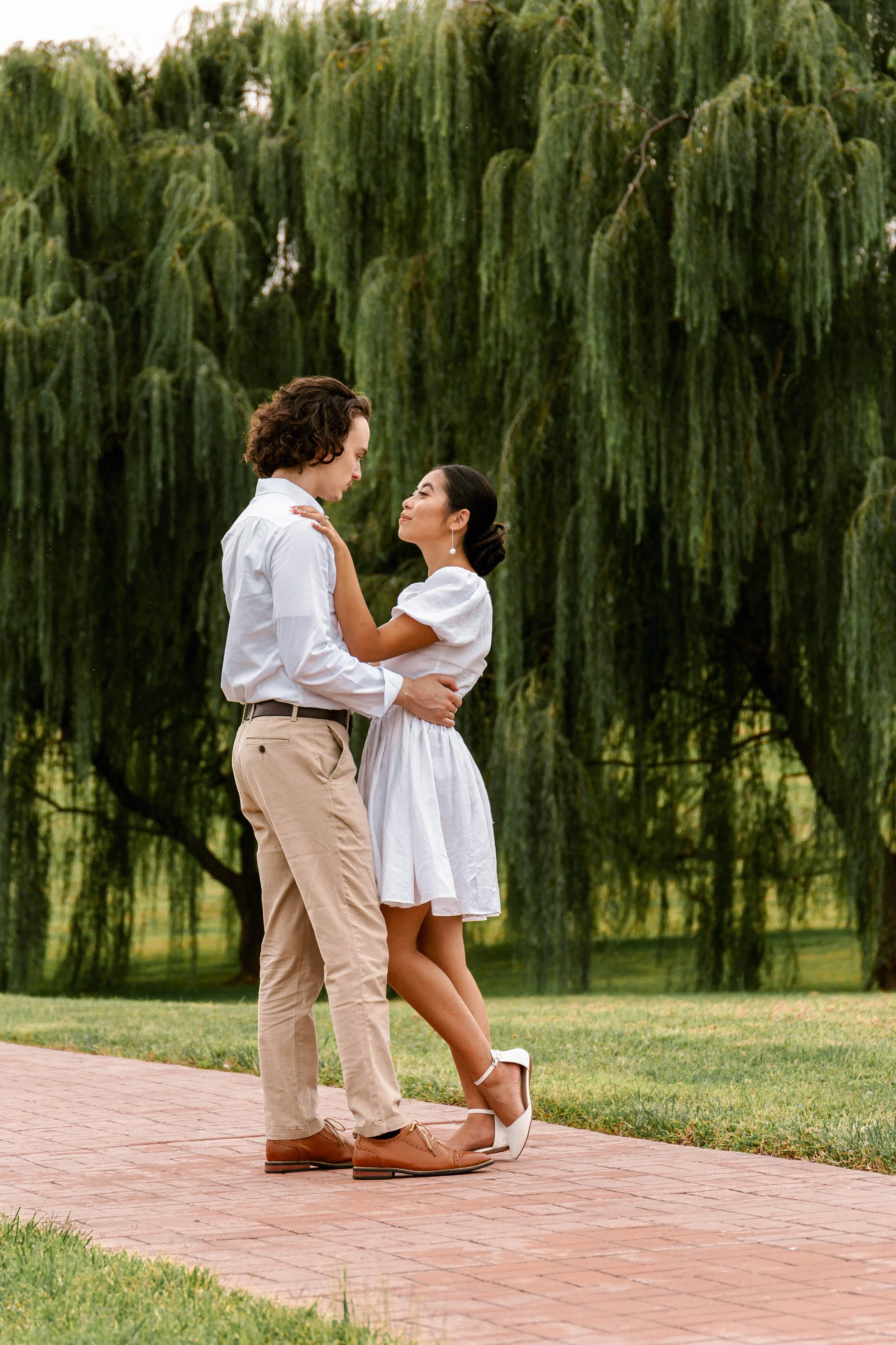 Couple embracing on a path in a park, surrounded by lush greenery and large willow trees.