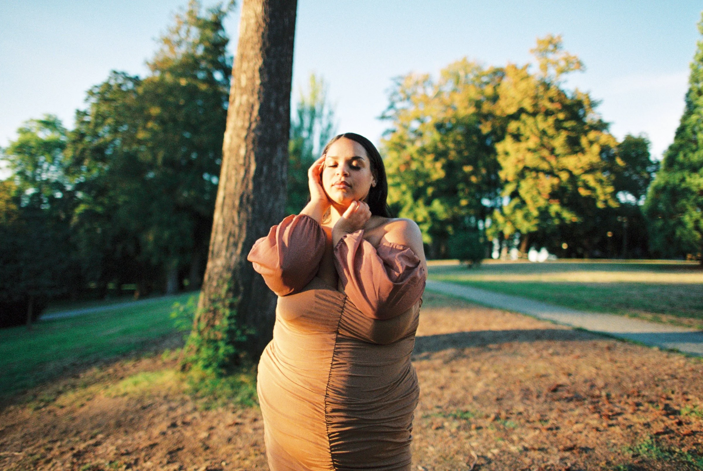 A woman with long dark hair standing in a park with tall trees and a walking path, her eyes closed, her hands gently touching her face and neck, wearing a beige dress with puffed sleeves.