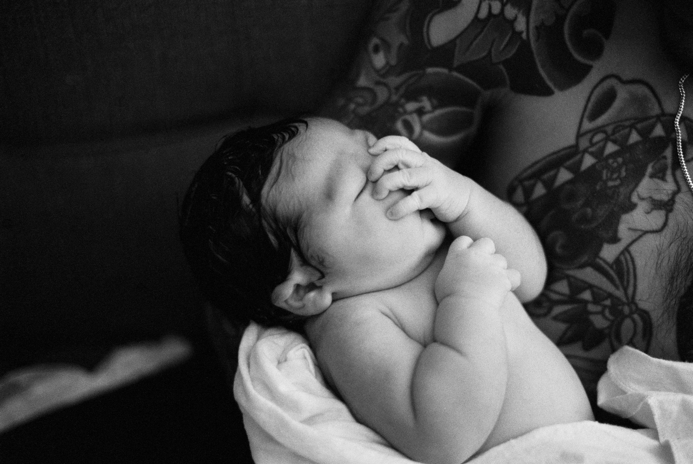 A newborn baby lying on a person's chest with tattoos, sleeping with one hand over their face in black and white photo.