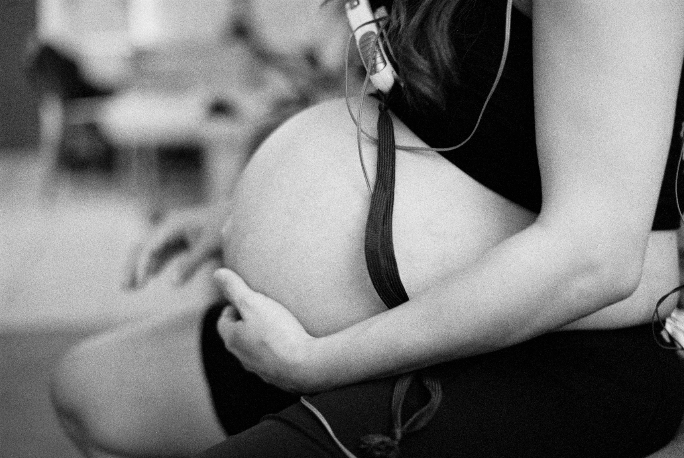 Black and white photo of a pregnant woman holding her belly, with earphones hanging from her neck, sitting in an indoor setting.
