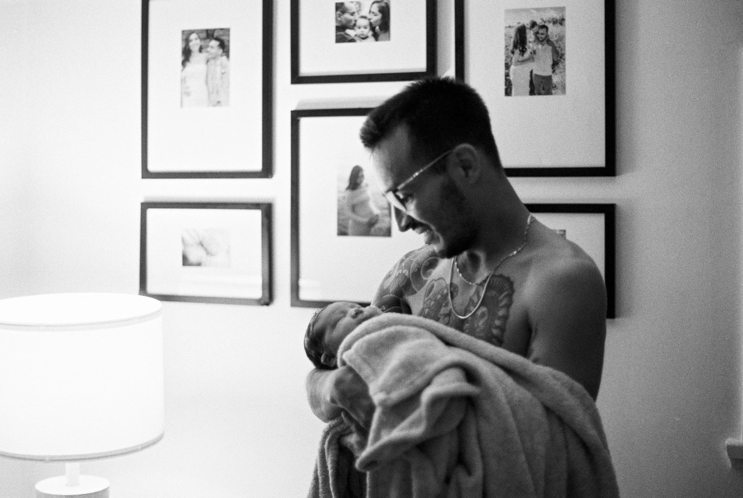A shirtless man with glasses and tattoos holding a newborn wrapped in a blanket, smiling at the baby in a room with framed family photos on the wall and a table lamp.
