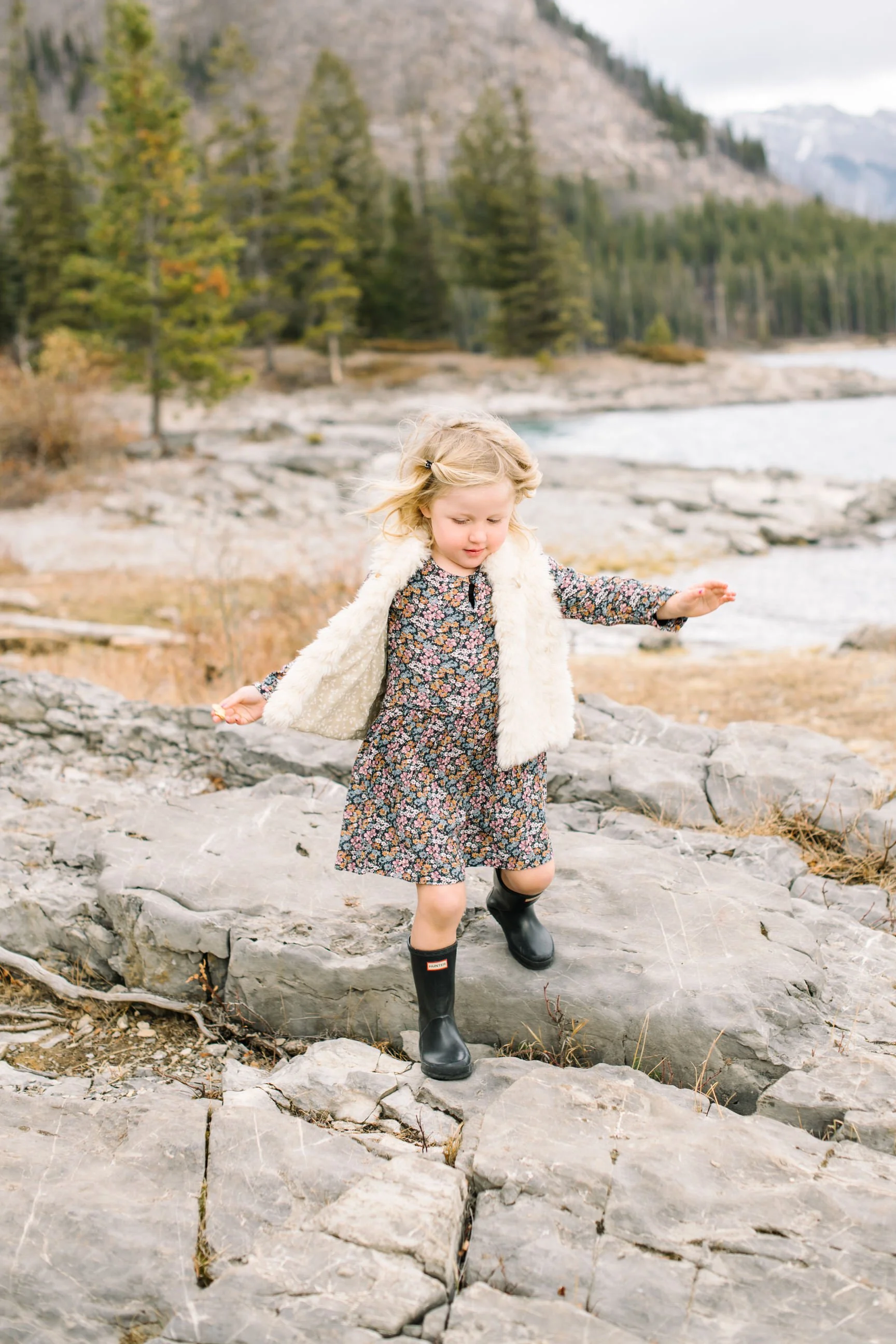 A young girl with blond hair walking on rocky terrain near a river, surrounded by trees and mountains in the background, wearing a floral dress, black rubber boots, and a white furry vest.