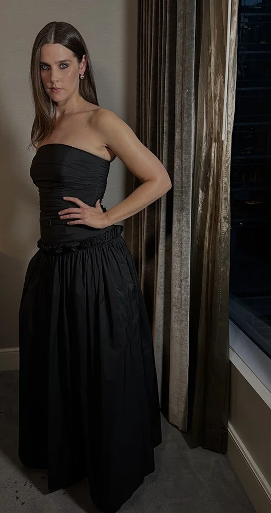 A woman with long brown hair wearing a strapless black dress standing indoors near a window with curtains at night.