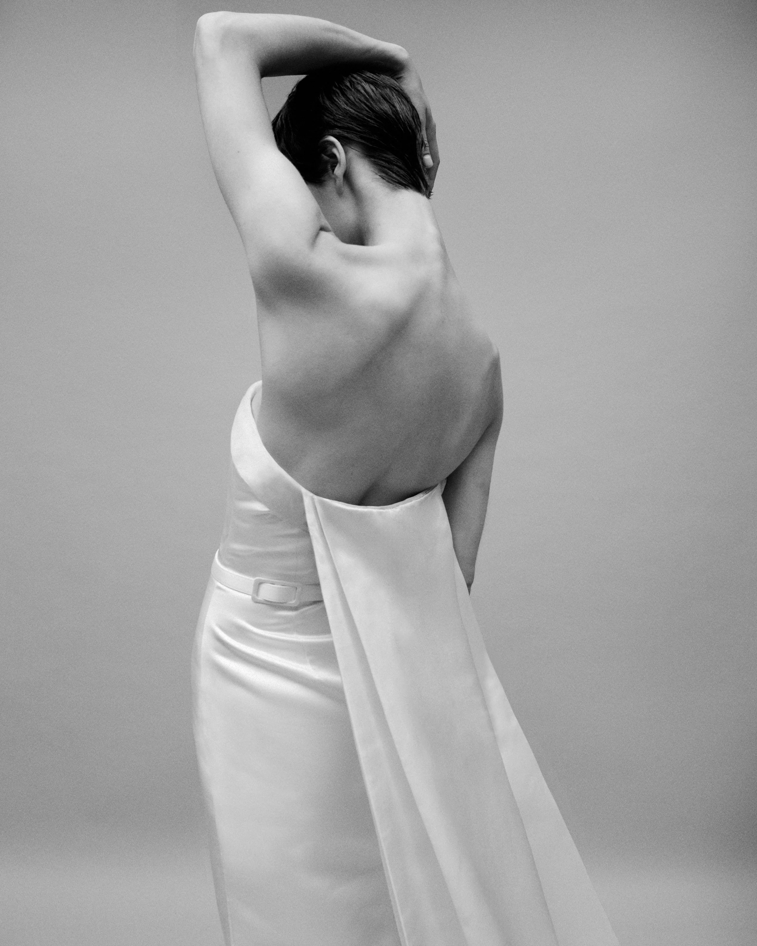 Black and white photo of a woman with her back to the camera, topless, with one hand behind her head and wearing a long, high-waisted skirt.