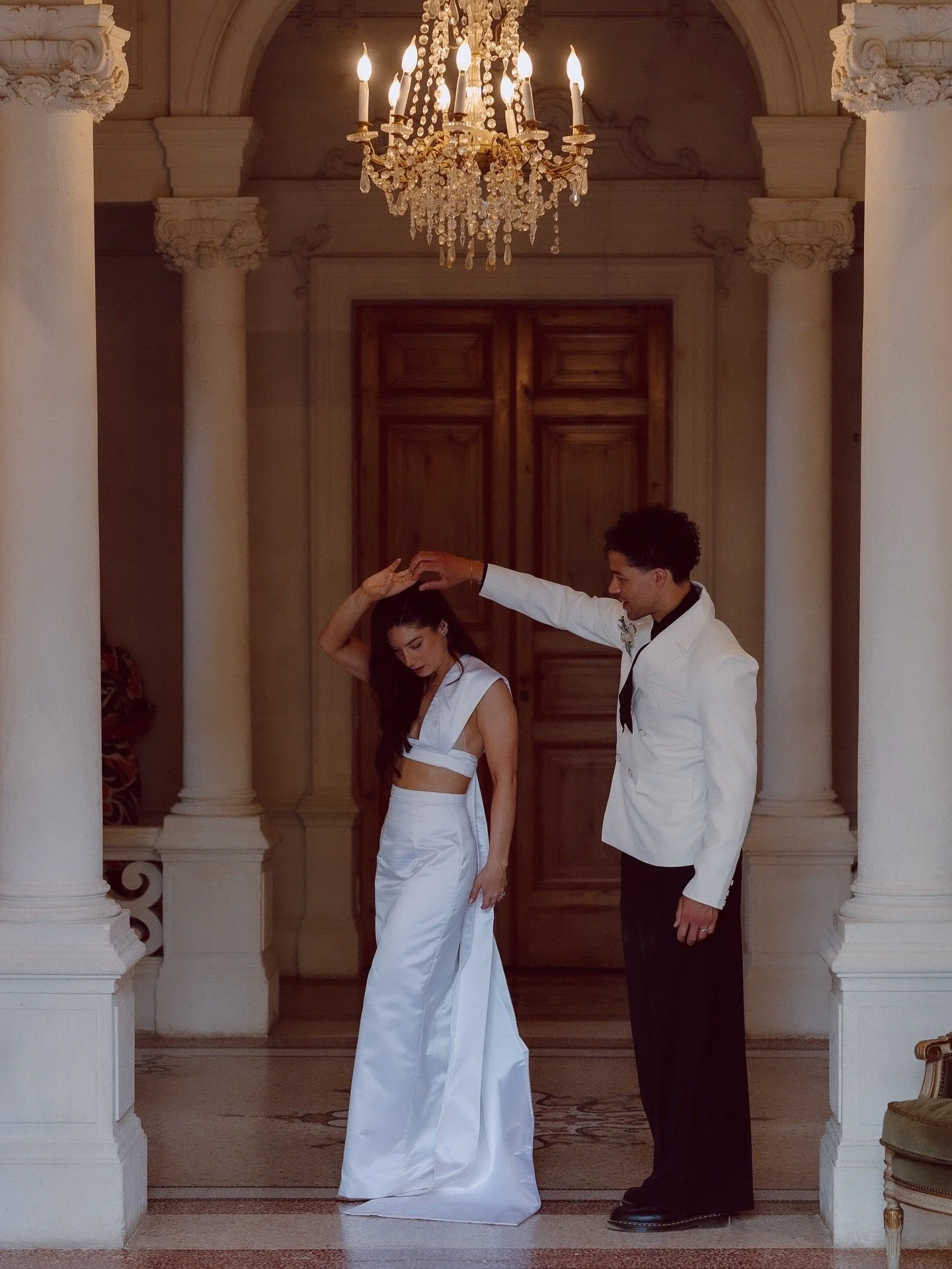 A man in a white tuxedo is dancing with a woman in a white wedding dress under a chandelier in a grand room with columns and wooden double doors.