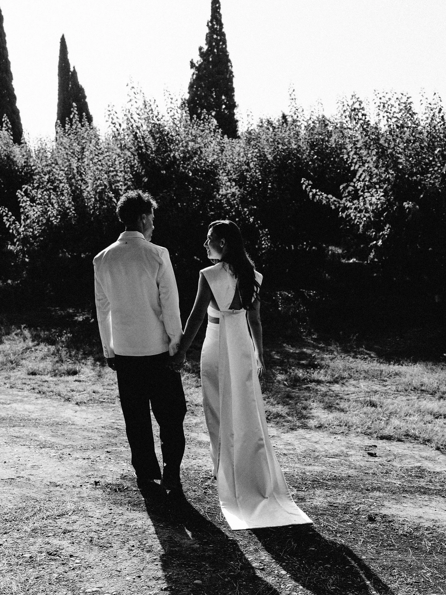 A couple dressed in formal attire walking hand in hand outdoors on a dirt path, with trees and bushes in the background, captured in black and white.