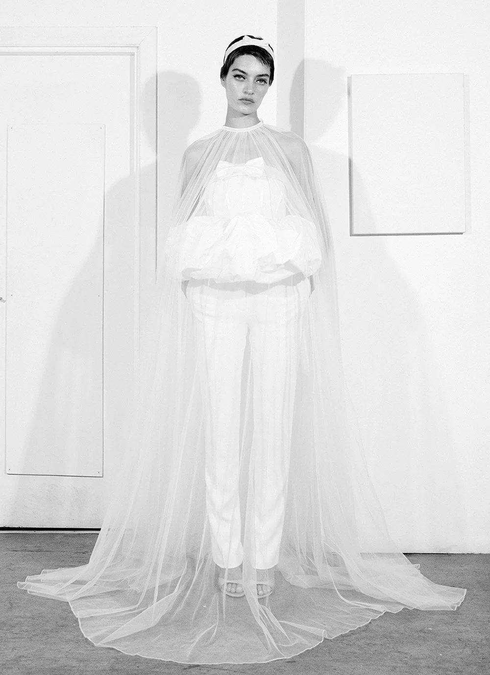Black and white photo of a woman wearing an elegant, flowing white gown with a sheer cape and a headband, standing in a minimalist room.
